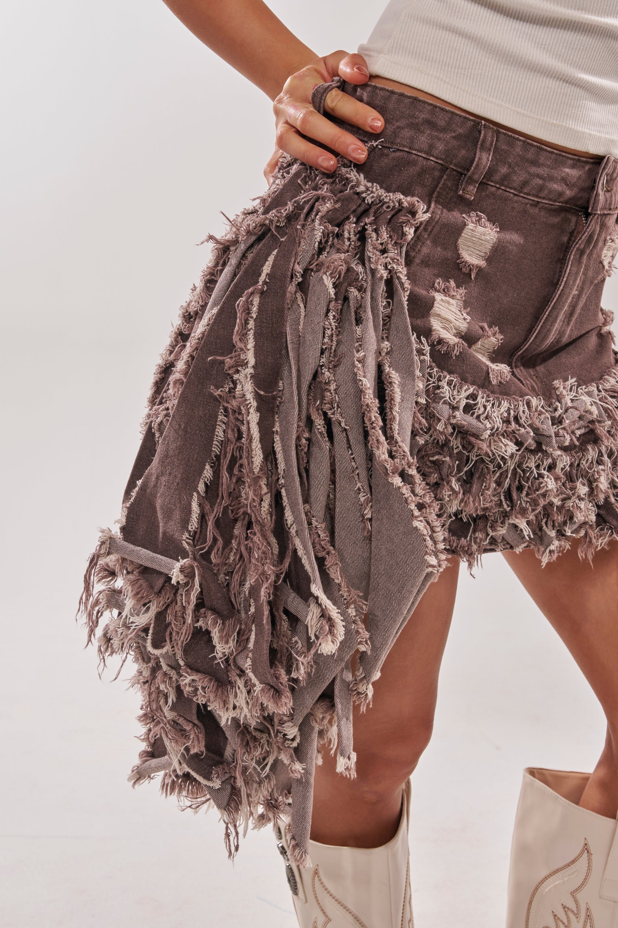 Wearing the TO THE RODEO WE GO SKIRT featuring distressed fringe and frayed details, paired with a white sleeveless top and white cowboy boots, one hand rests on their hip. 9