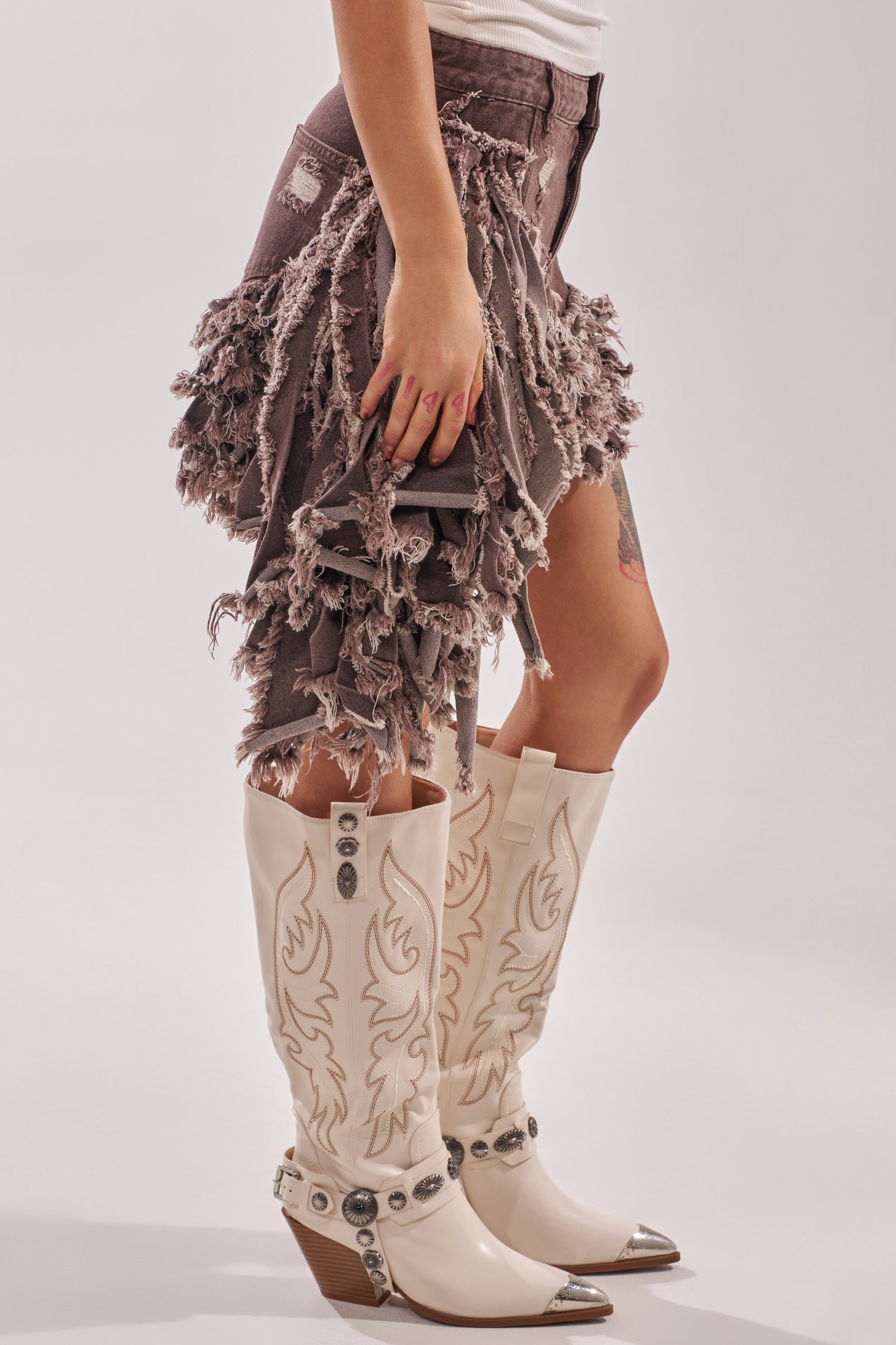 Wearing the TO THE RODEO WE GO SKIRT and purple and white cowboy boots with decorative stitching and metal details, a person stands against a plain background. 3