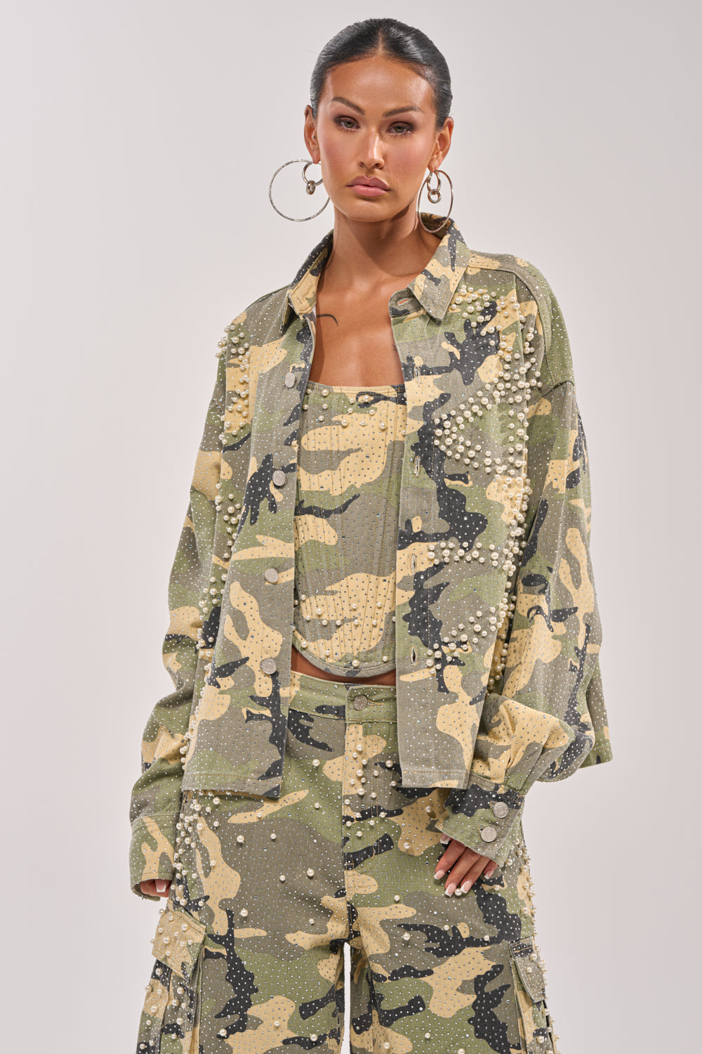 A woman stands confidently in the EMBELLISHED CAMO BUTTON DOWN with matching pants, hands in pockets. She wears hoop earrings and a sleek hairstyle, exuding a modern street-luxe vibe against a plain backdrop. 0