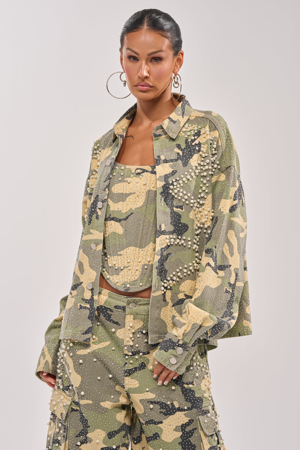 A woman wears the EMBELLISHED CAMO BUTTON DOWN, her hair tied back and hoop earrings shining. She poses with a neutral expression against a plain background. 2