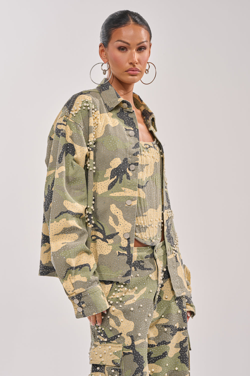 A woman stands facing forward against a plain light background, wearing the EMBELLISHED CAMO BUTTON DOWN with pearl accents, hoop earrings, and her hair pulled back. 3