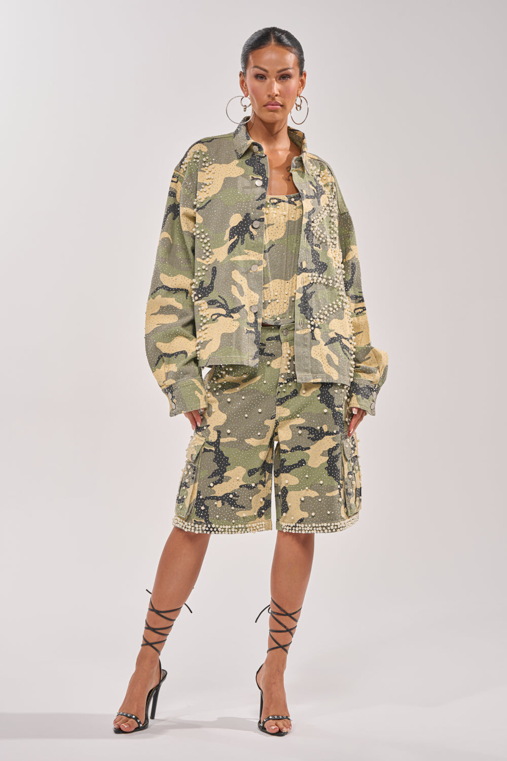 A woman exudes confidence in the EMBELLISHED CAMO BUTTON DOWN, paired with a matching top and knee-length shorts. Black strappy heels and large hoop earrings complete her bold, fashion-forward ensemble. 7
