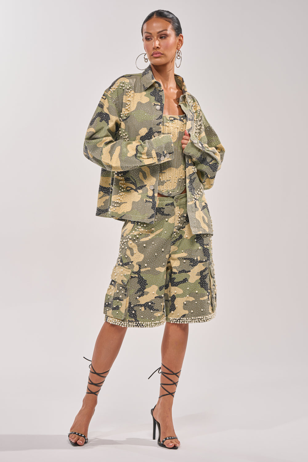 A woman models the EMBELLISHED CAMO BUTTON DOWN with matching camo knee-length shorts, a coordinating top, and strappy black heels. Large hoop earrings and slicked-back hair complete her chic, fashion-forward look. 8