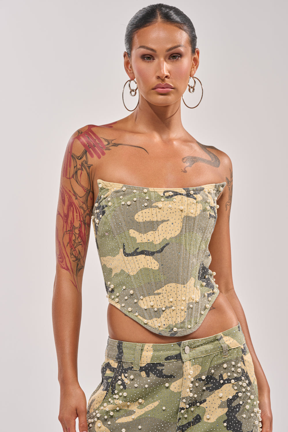 Model wears the EMBELLISHED CAMO CORSET, a strapless design with pearl rhinestones, paired with matching camo pants. Hoop earrings, visible tattoos, and a slicked-back hairstyle enhance her edgy streetwear vibe. 1