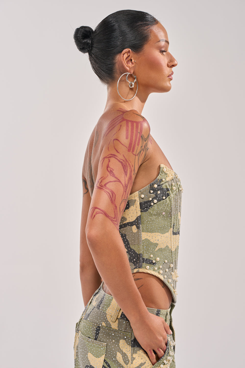 A woman with slicked-back hair, hoop earrings, and a bold arm tattoo models the EMBELLISHED CAMO CORSET in profile against a plain background, creating an edgy streetwear statement. 3