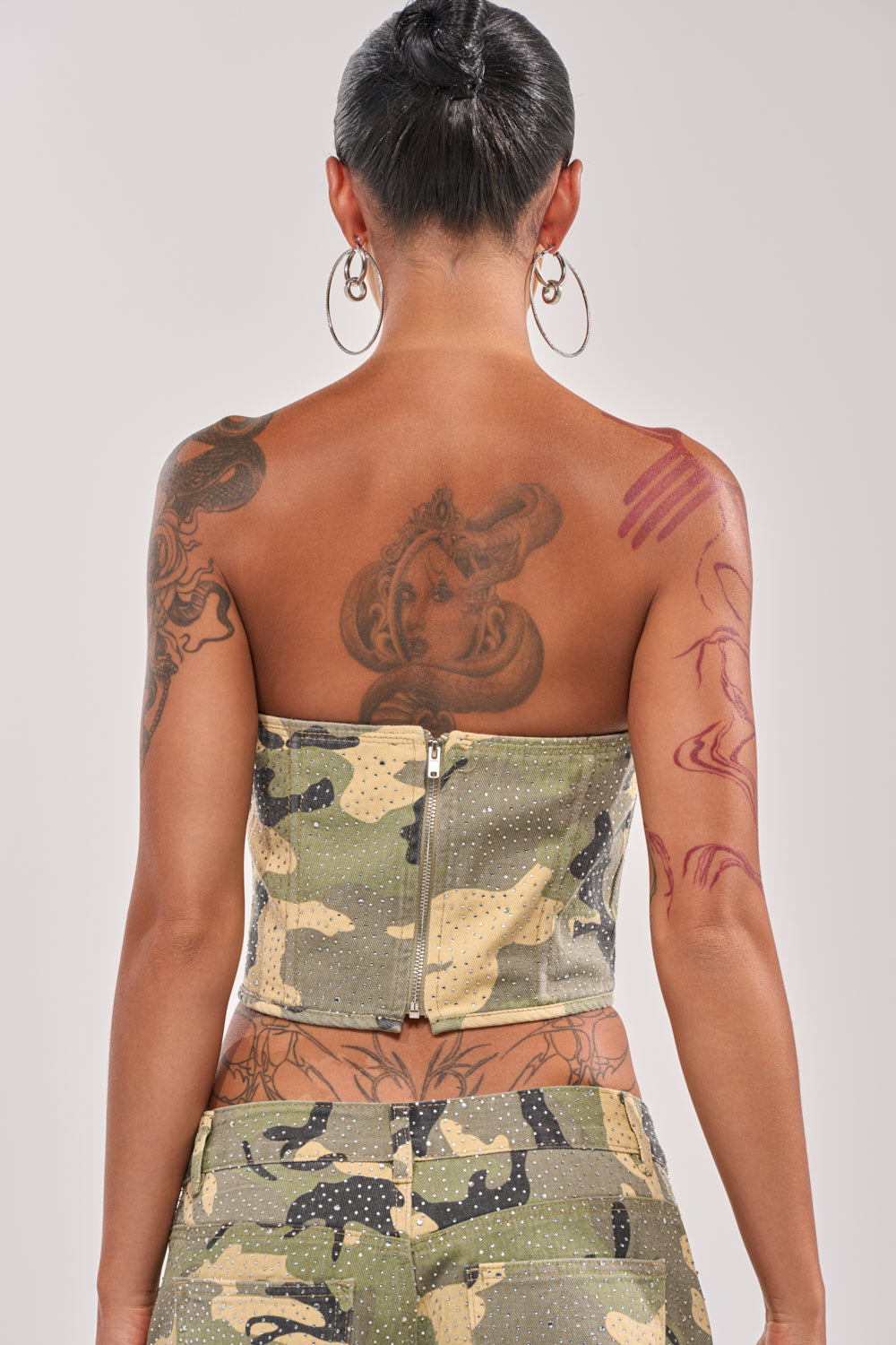 A person with short hair and tattoos is shown from the back wearing the EMBELLISHED CAMO CORSET with matching pants. Large hoop earrings complete the streetwear-inspired look against a plain background. 4