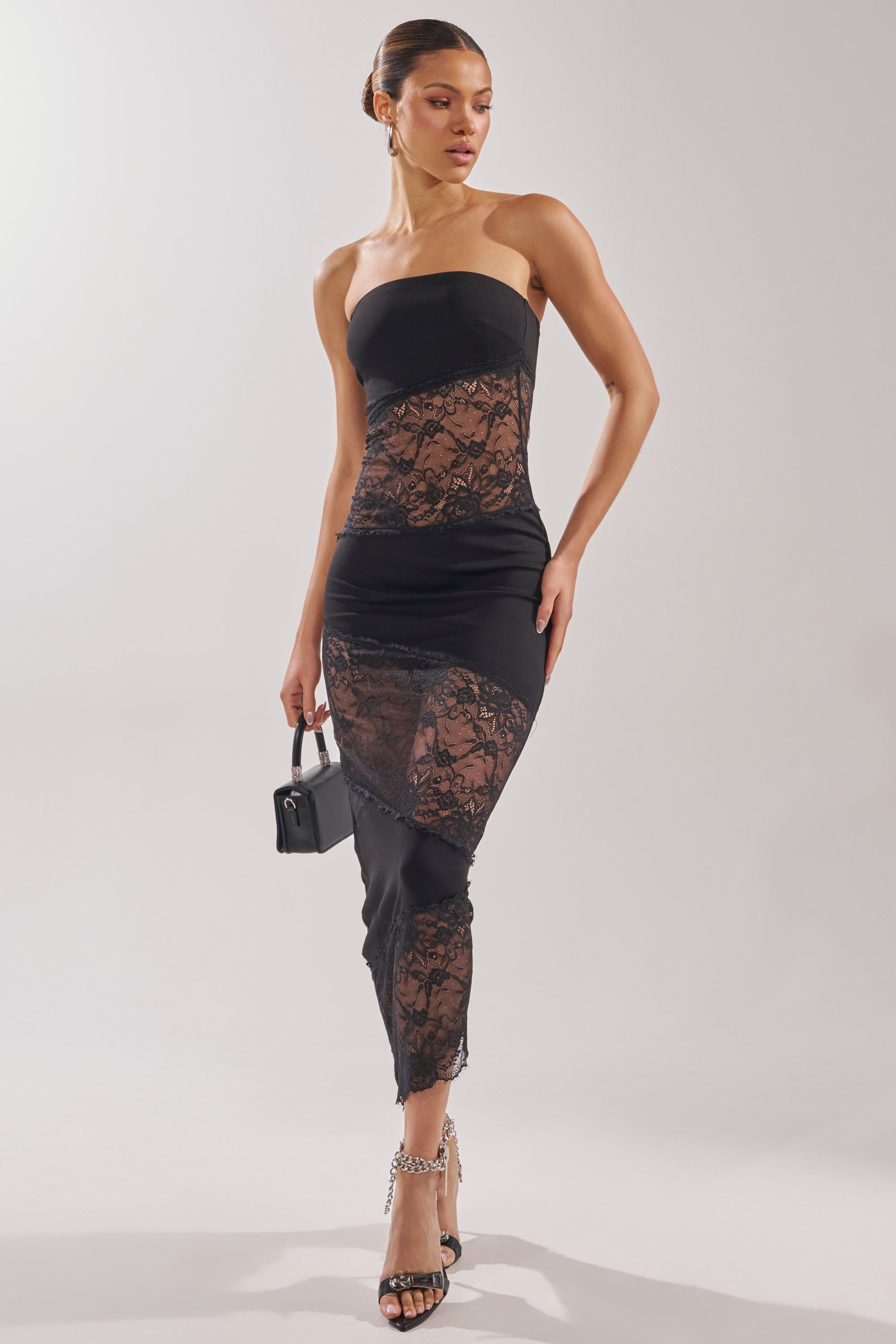 A woman poses in the ILL SHOW YOU LACE MAXI DRESS, featuring sheer lace paneling. She holds a small black handbag, wears black high-heeled sandals, and her hair is in a sleek bun. The background is plain and light-colored.