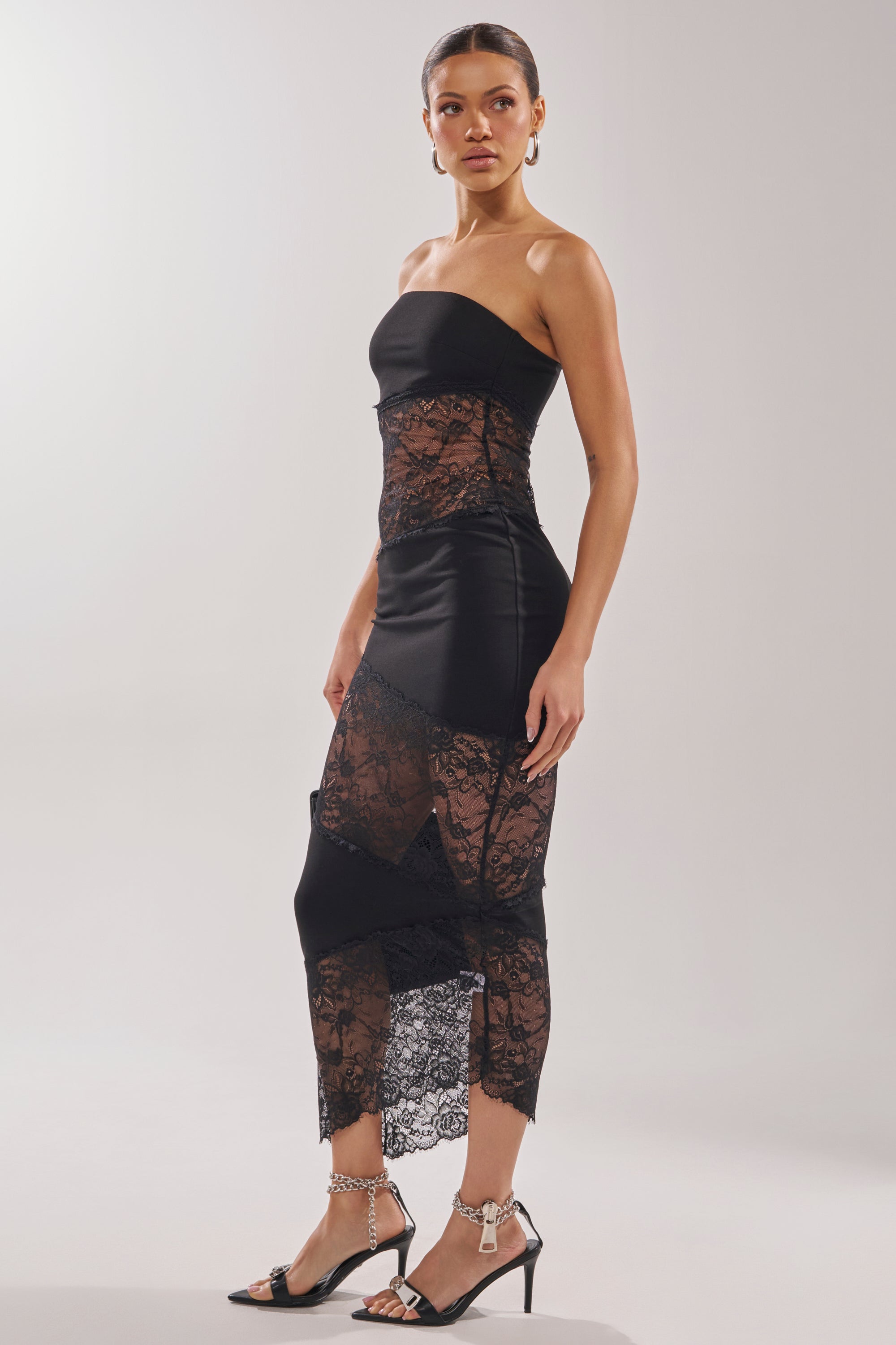 A woman wears the ILL SHOW YOU LACE MAXI DRESS, featuring strapless styling and sheer lace panels, paired with black ankle-strap sandals, standing against a plain light background.