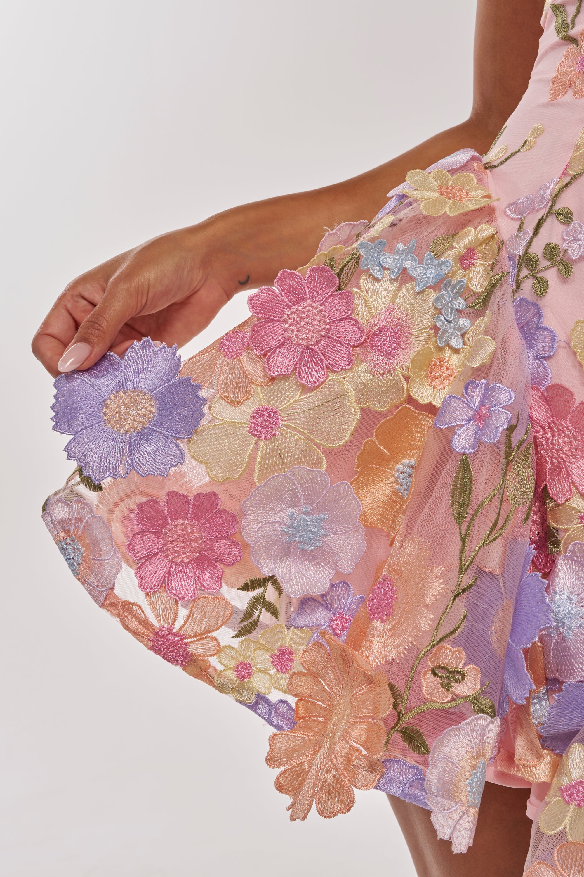 A close-up of a hand holding the edge of the SHE WAS A FAIRY MINI DRESS, featuring a sheer tulle overlay decorated with embroidered flowers in pink, purple, peach, and yellow. 10