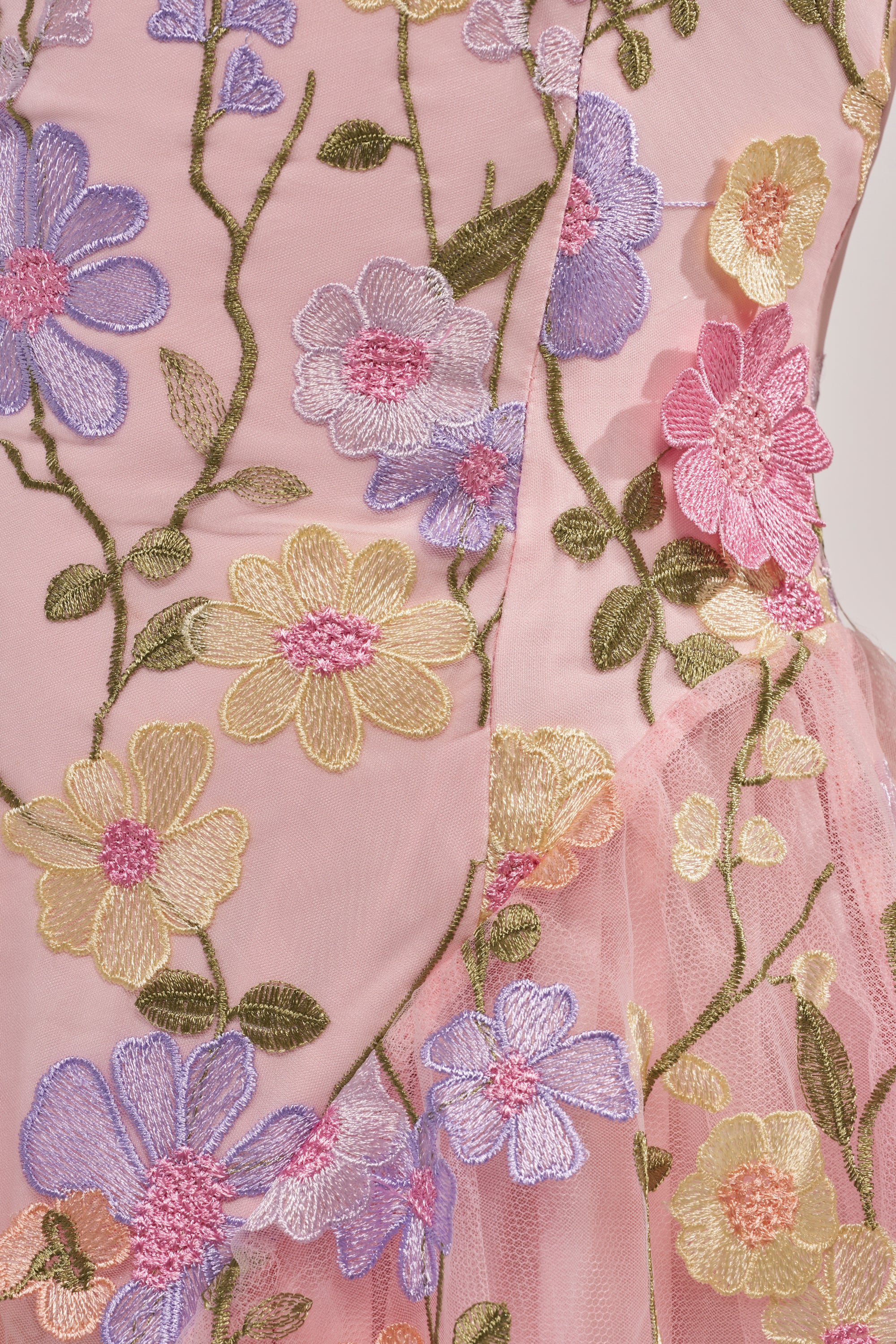 Close-up of pink fabric from the SHE WAS A FAIRY MINI DRESS, featuring intricate embroidered pastel flowers—purple, yellow, and pink blooms with green stems and leaves. The detailed embroidery adds texture to this summery piece. 9