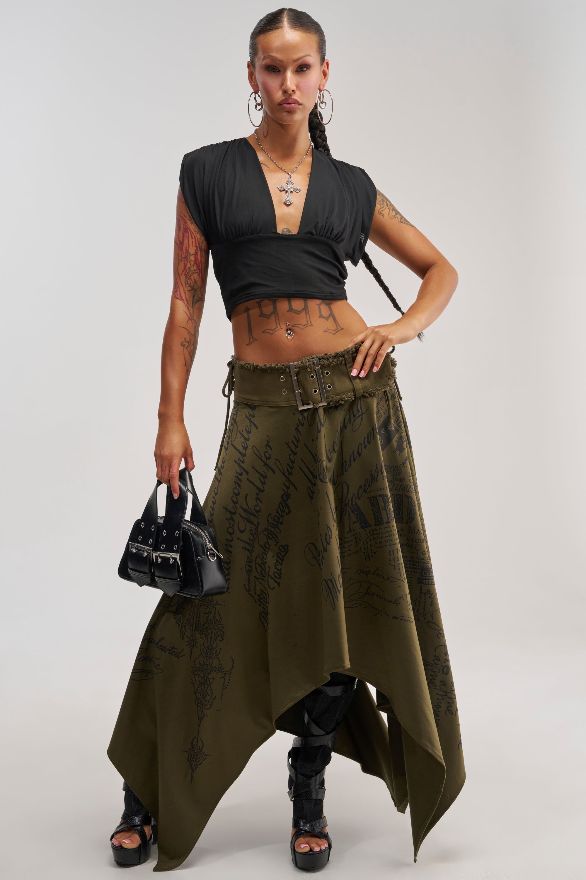 Woman with long braids wearing a black crop top, the NONE OF YOUR CONCERN MIDI SKIRT—an olive handkerchief skirt with bold black typography print—black heeled shoes, hoop earrings, and holding a small black handbag.