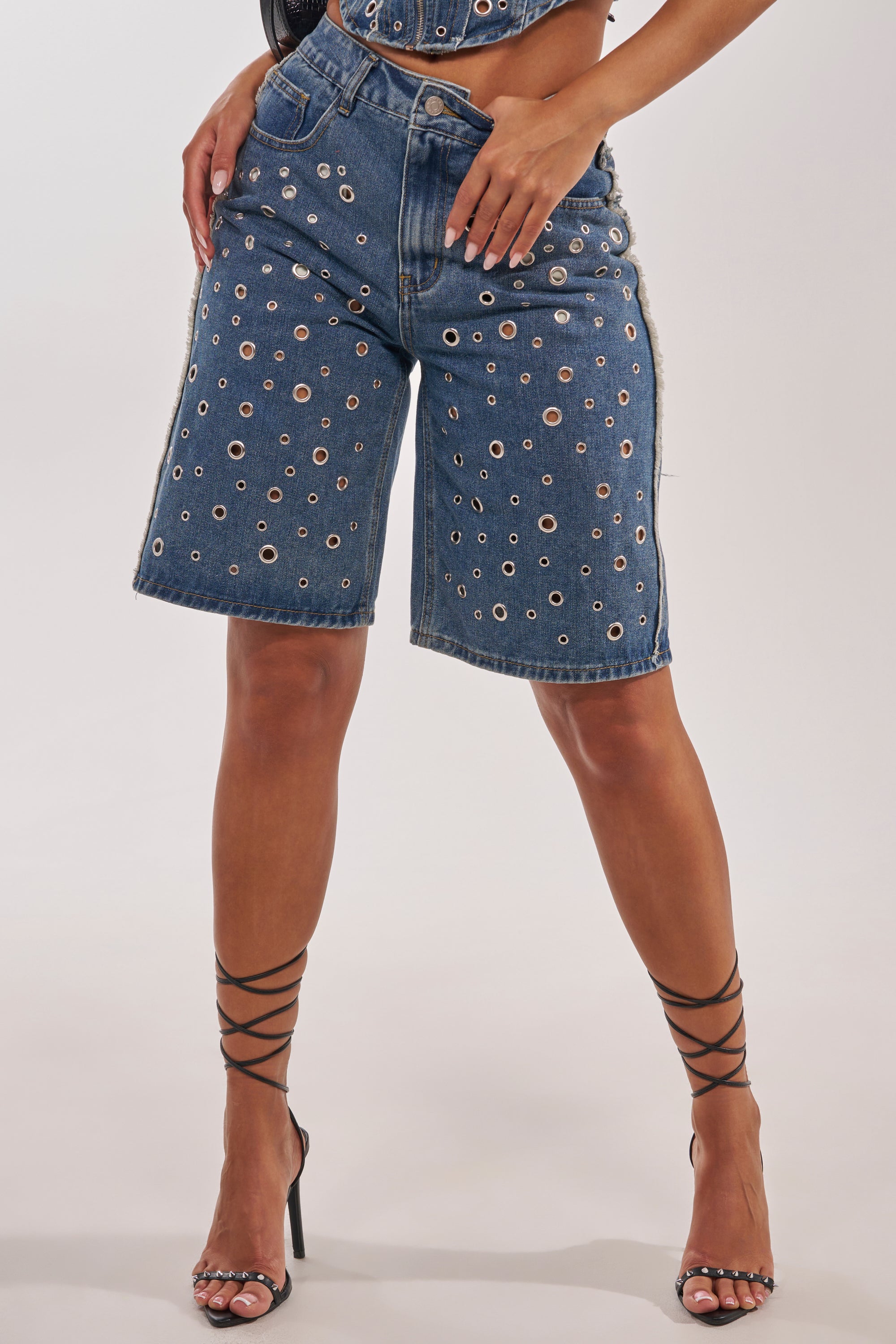 A person stands against a plain background wearing the TAYLOR DENIM BERMUDA WITH GROMMET, high-rise blue denim Bermuda shorts featuring silver grommet hardware, paired with black crisscross strap high-heeled sandals. 1