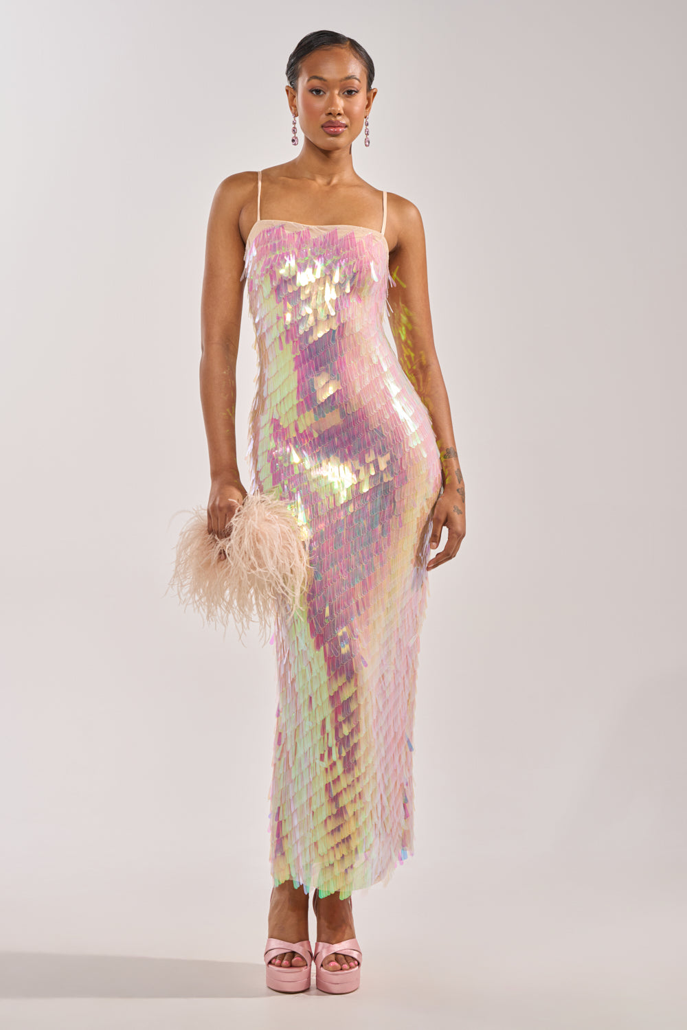 A woman wears the ALWAYS SHINING SEQUIN MAXI DRESS with spaghetti straps, styled with pink platform heels, pink earrings, and a pale fluffy clutch. Her hair is neatly styled back against a plain light background. 0