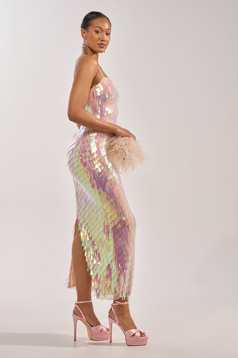 A woman in the ALWAYS SHINING SEQUIN MAXI DRESS poses sideways, wearing pink platform heels and holding a pale pink feathered clutch, with her hair pulled back against a plain light background—a standout special occasion look. 2