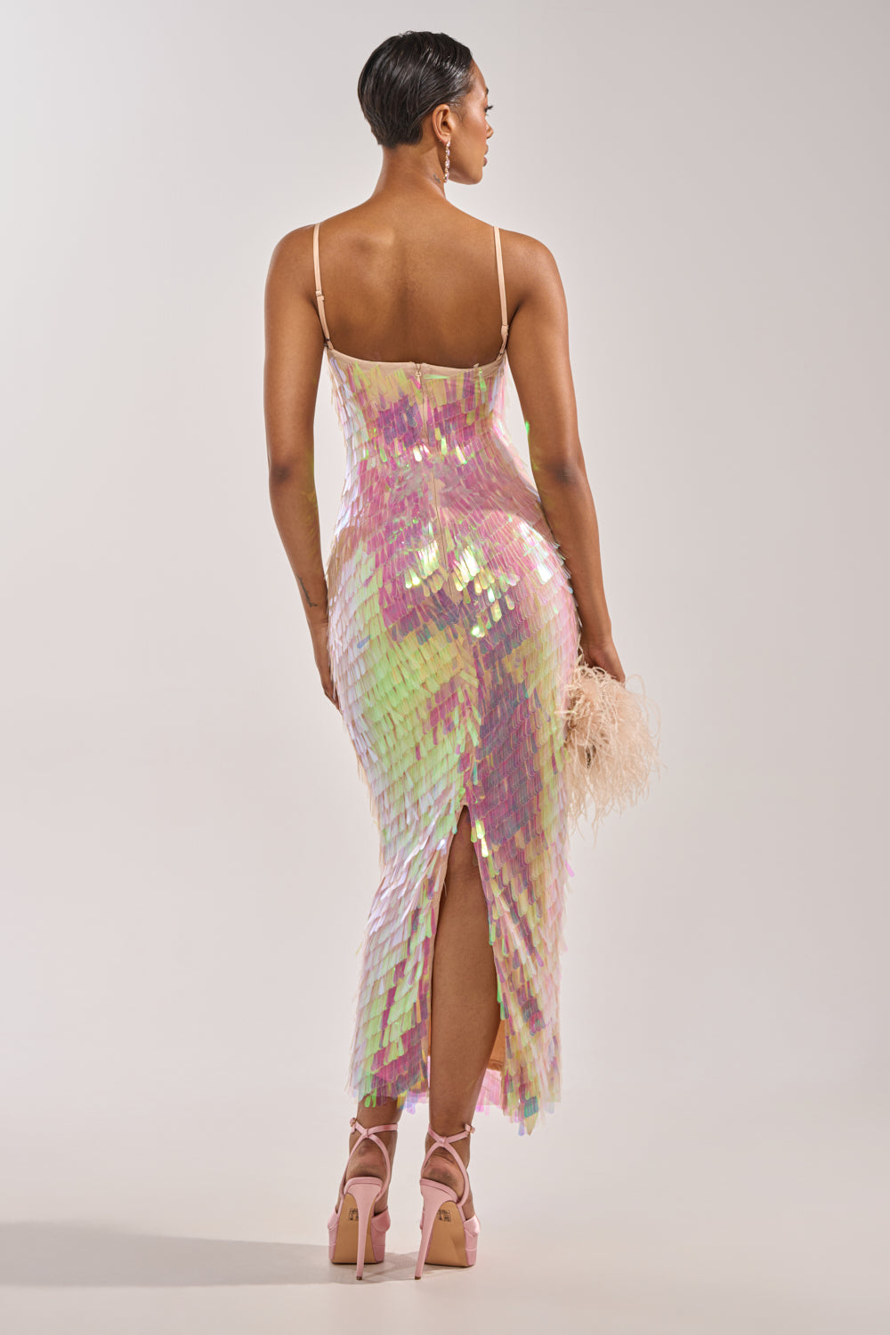 A woman faces away in the ALWAYS SHINING SEQUIN MAXI DRESS, featuring iridescent sequins, thin straps, and a back slit. She pairs it with a feathered clutch, high pink platform heels, and chic short hair—ideal for special occasions. 3
