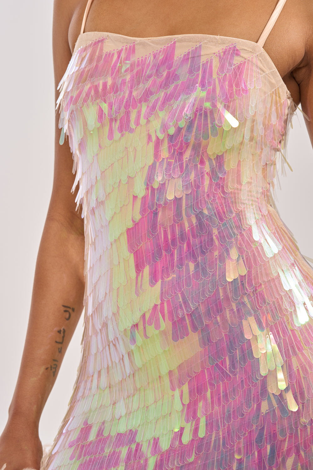 A close-up of a person wearing the ALWAYS SHINING SEQUIN MAXI DRESS, a sleeveless iridescent sequin maxi with layered pink, yellow, and green accents. Their arm, featuring a small tattoo, is visible. 4