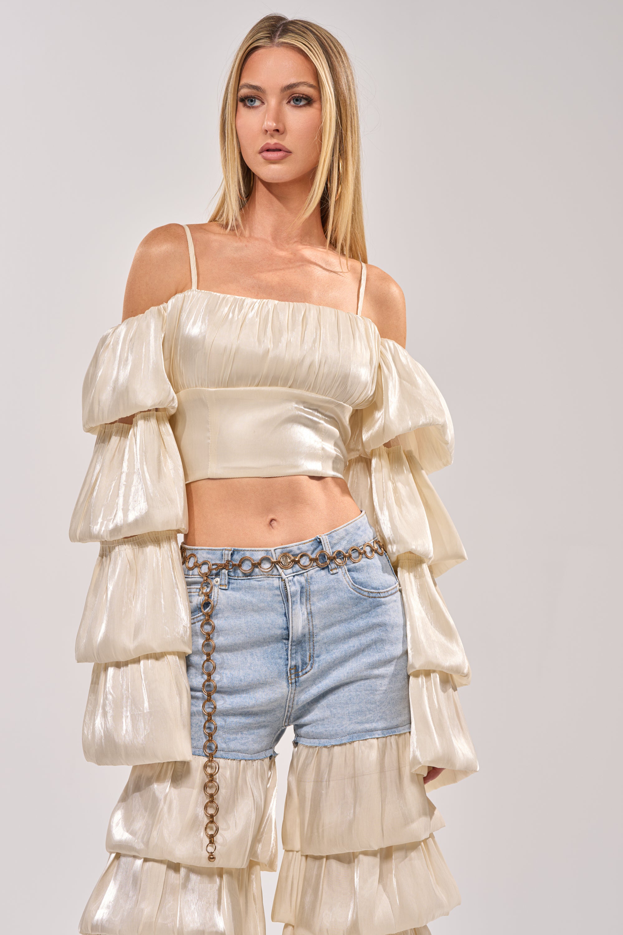 A woman with straight blonde hair wears the BOUNCIN AROUND BLOUSE—an ivory off-shoulder crop top with tiered sleeves—styled with light blue denim shorts and a chain belt, standing against a plain background. 0