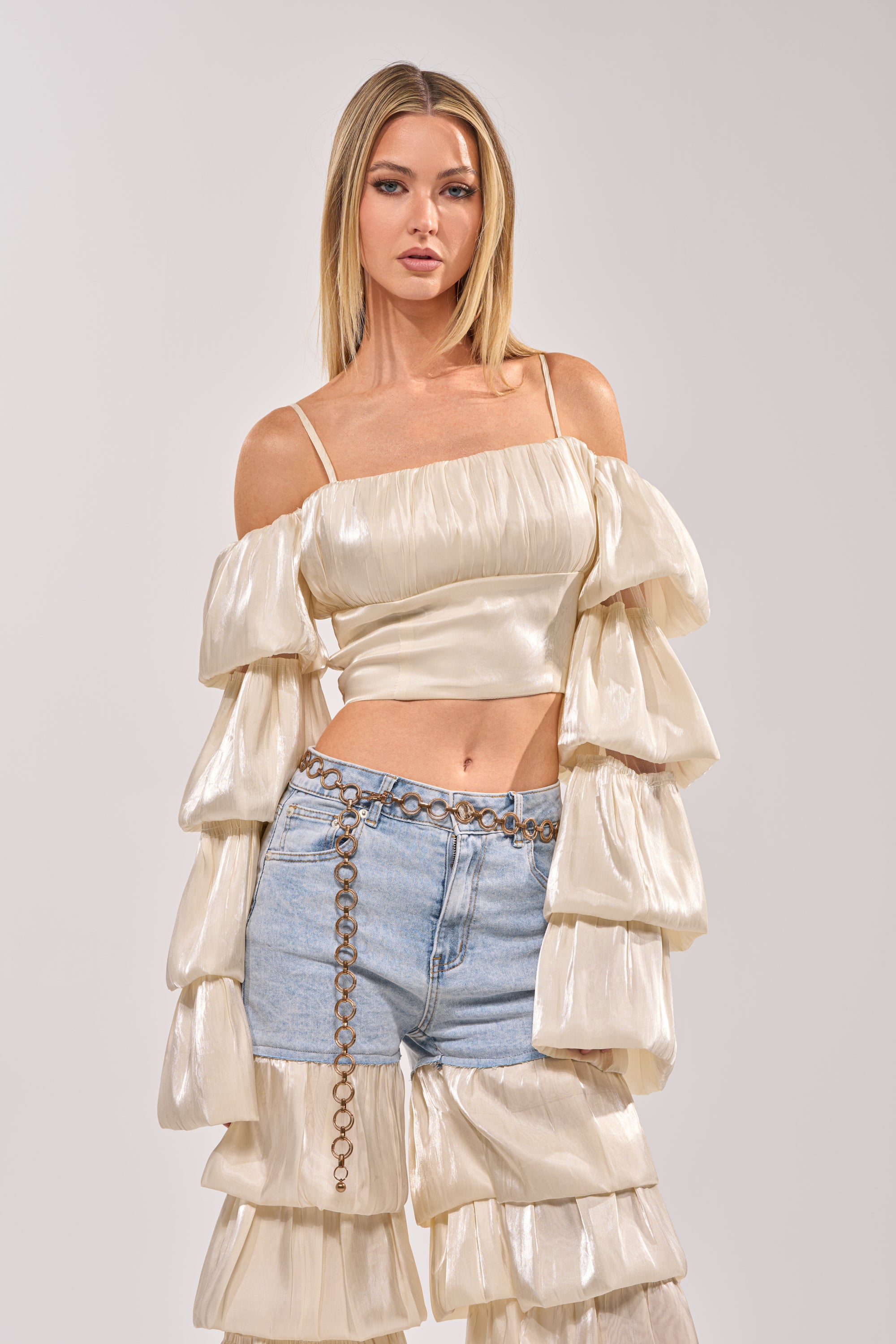 A woman models the BOUNCIN AROUND BLOUSE, an ivory off-shoulder crop top with tiered sleeves and ruffles, paired with light blue denim jeans and a chain belt. She has straight blonde hair and a neutral expression. 1