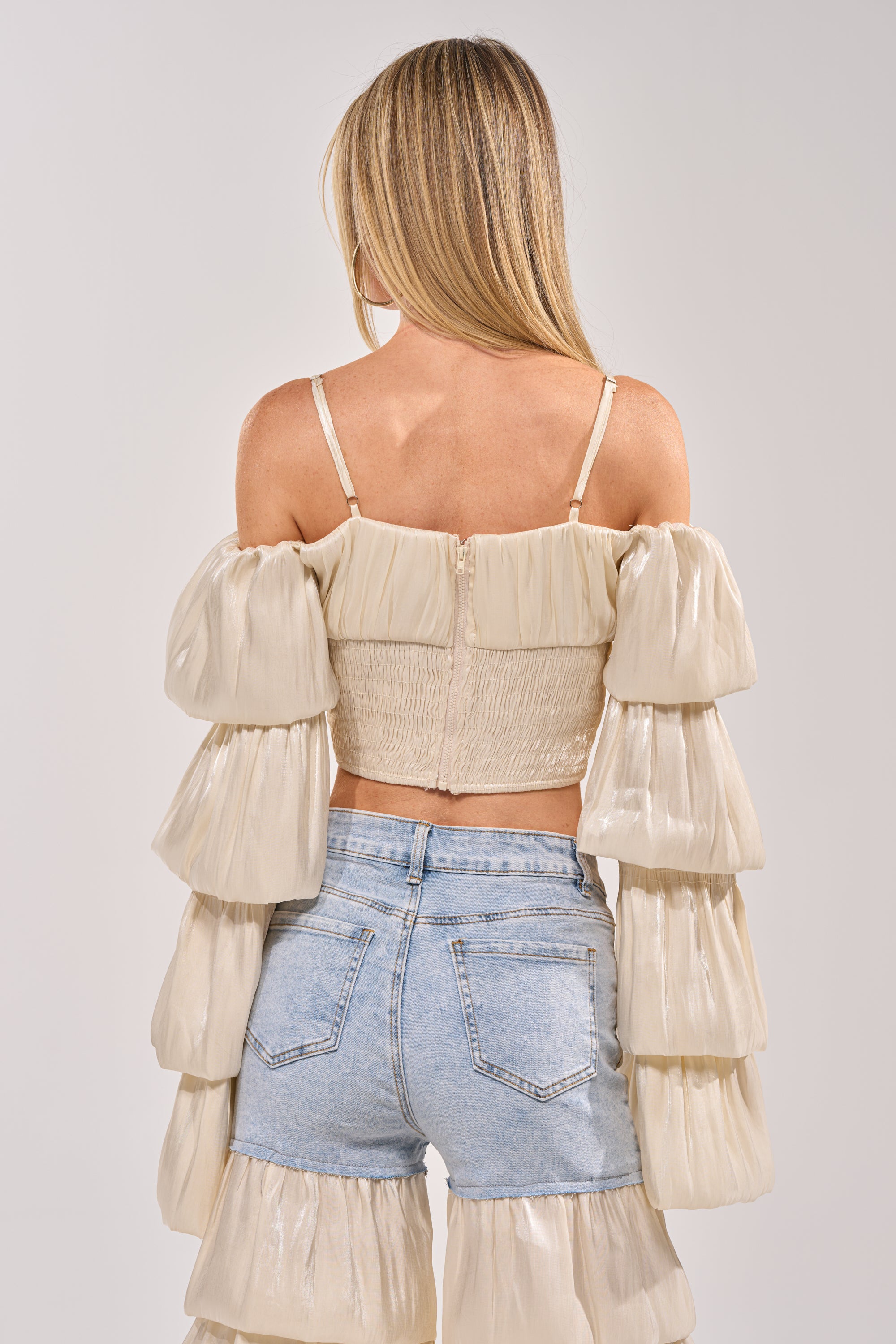 A woman with long blonde hair, seen from the back, wears the BOUNCIN AROUND BLOUSE—an ivory off-shoulder crop top with tiered ruffled sleeves, smocked back, and spaghetti straps—paired with light blue high-waisted denim shorts. 3