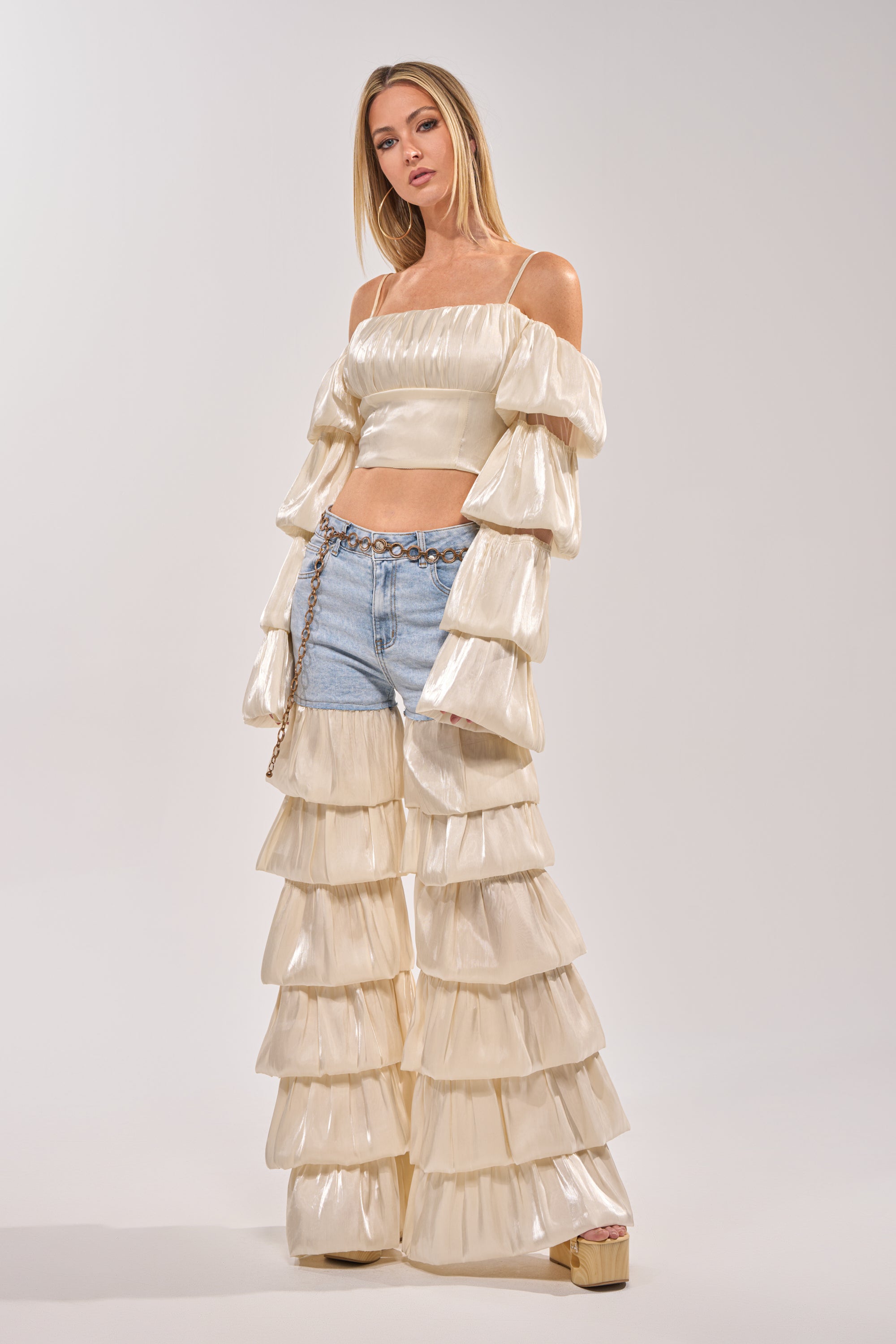 A woman poses in the BOUNCIN AROUND BLOUSE—an ivory off-shoulder crop top with matching tiered ruffle pants over denim shorts, styled with a chain belt, against a plain light gray background. 5