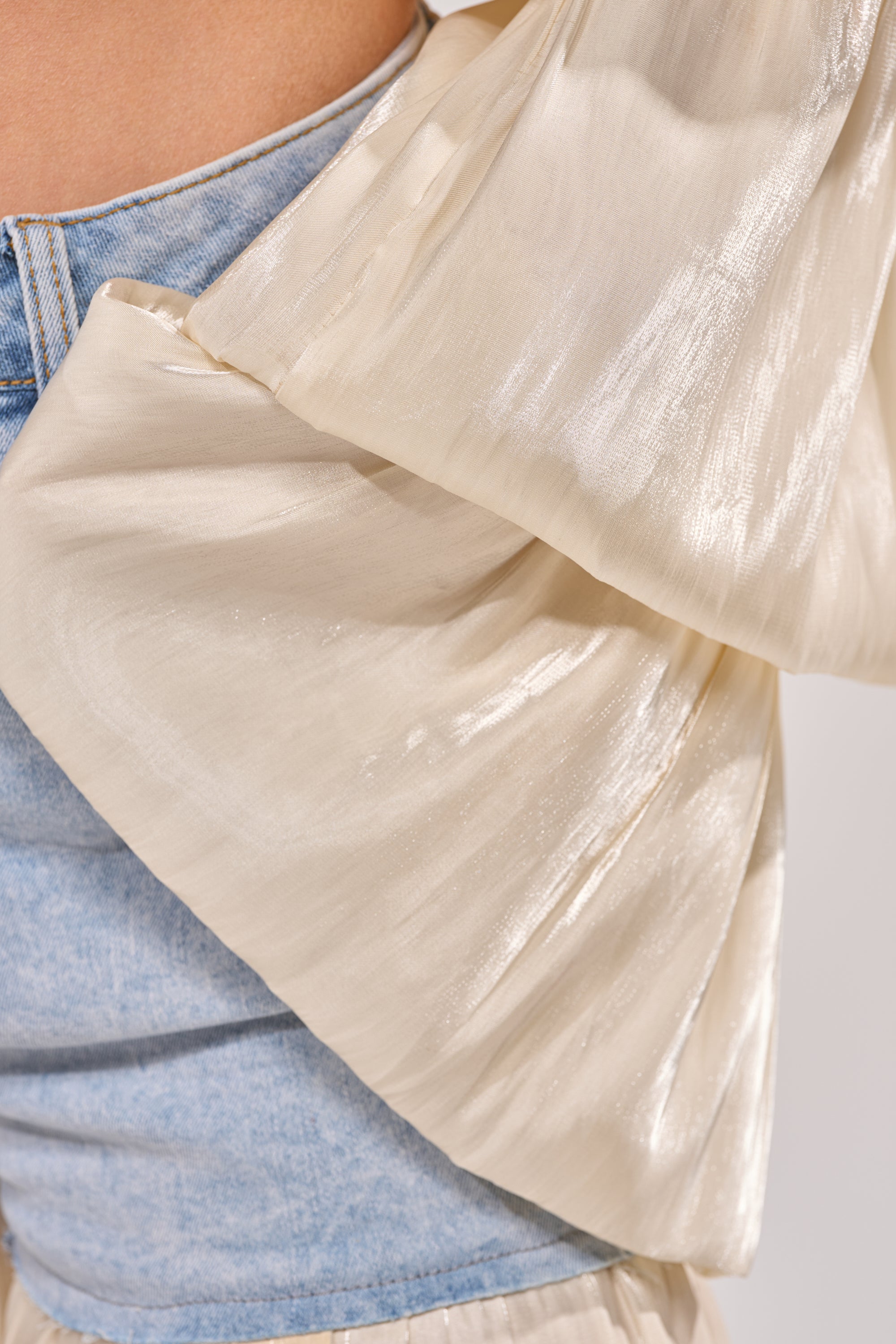 Close-up of a person wearing the BOUNCIN AROUND BLOUSE, a shimmery organza crop top with voluminous sleeves styled over light blue denim, revealing the shiny, textured fabric and denim underneath. 7