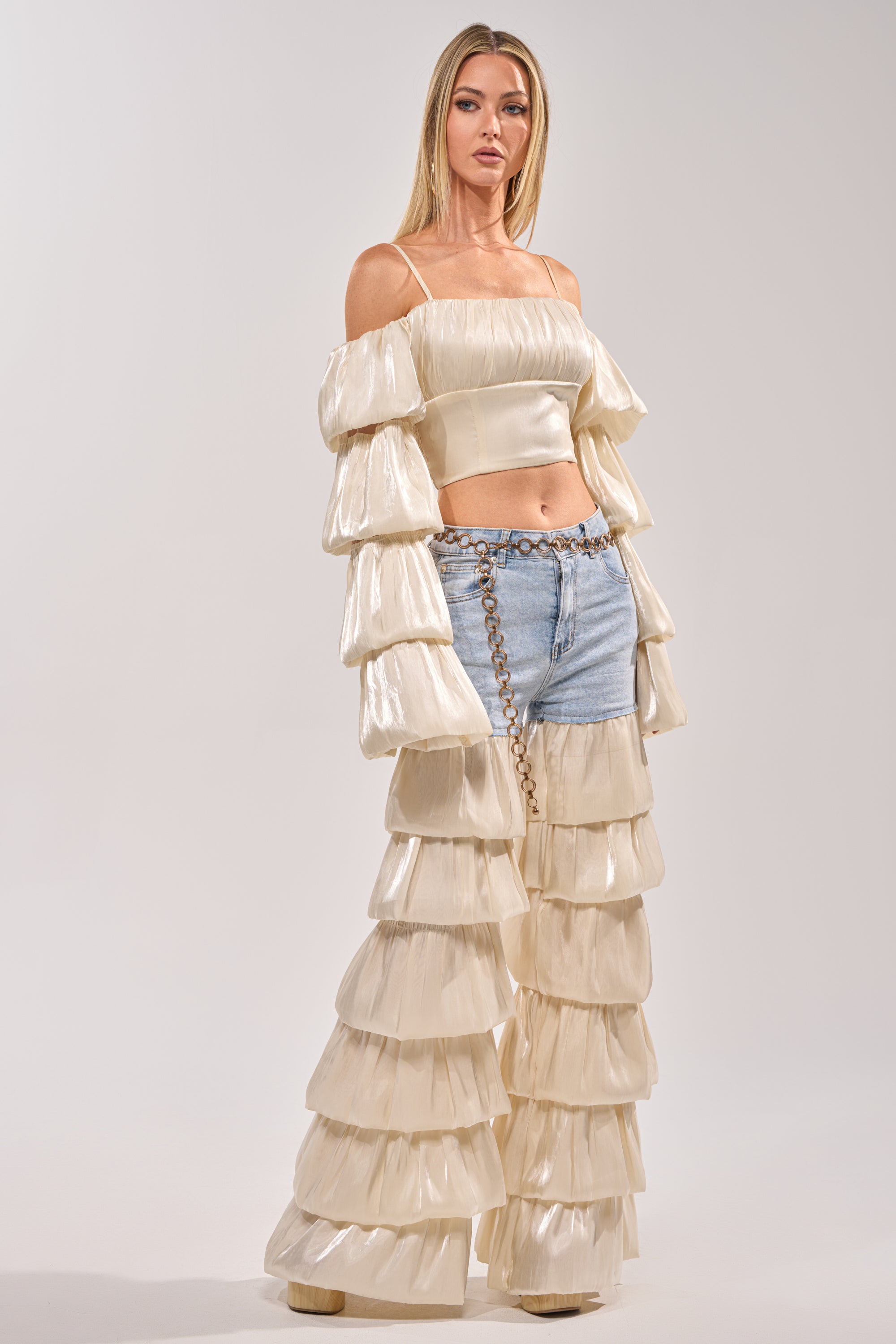 A woman with straight blonde hair and a neutral expression poses against a plain background, wearing the BOUNCIN AROUND DENIM RUFFLE PANT paired with a cream off-shoulder ruffle top, styled with a chain belt over statement denim. 0
