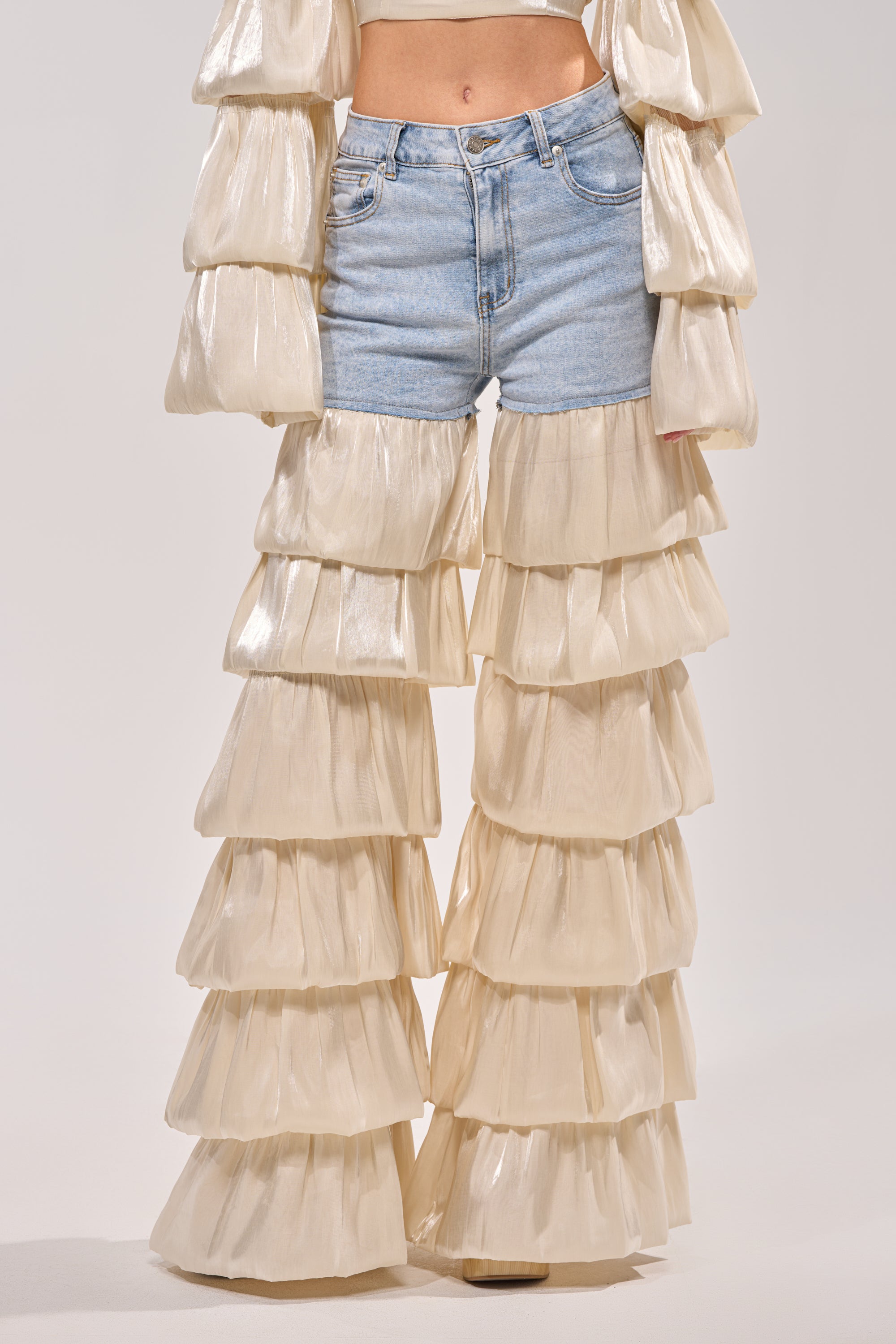 A person models the BOUNCIN AROUND DENIM RUFFLE PANT—light blue jeans with tiered organza ruffle bell-bottoms. The photo, shot on a neutral background, is cropped from chest to feet. 1