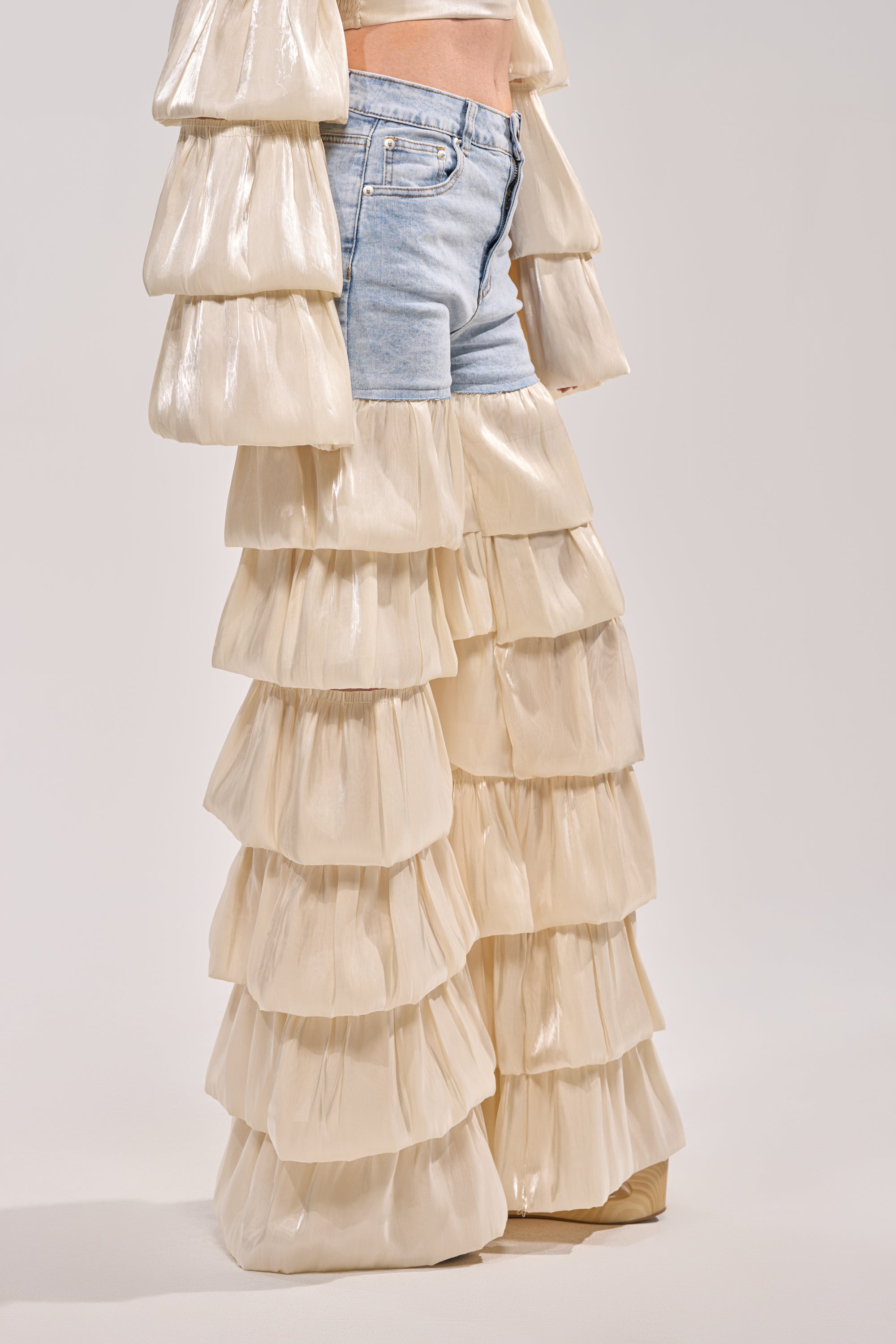 Wearing the BOUNCIN AROUND DENIM RUFFLE PANT layered over cream organza ruffle pants with matching sleeves, the standout jeans pop against a simple light backdrop. 2