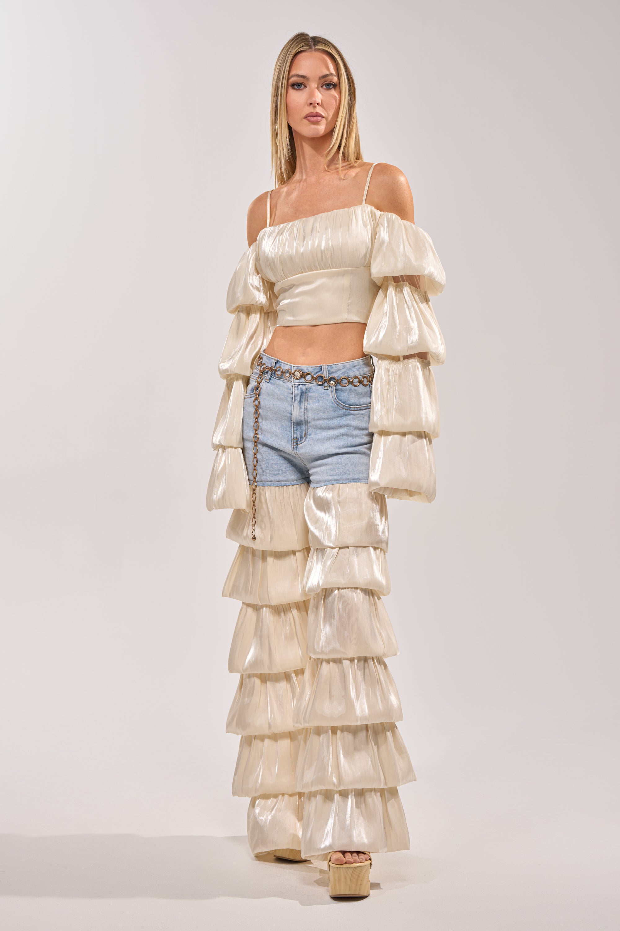 A woman wearing the BOUNCIN AROUND DENIM RUFFLE PANT, styled with a shiny cream off-shoulder top, chain belt, and platform sandals, poses against a light background. 5