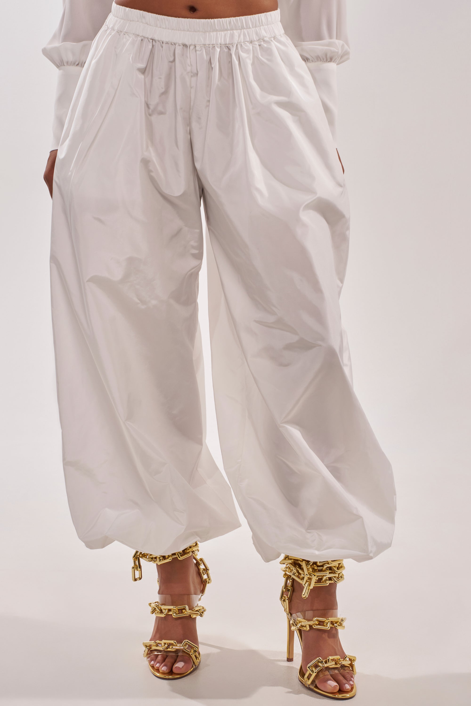 Someone stands against a plain background wearing the EFFORTLESS BALLOON PANT IN WHITE with an elasticized waistband. Their upper body is mostly out of frame, and they pair the pants with gold high-heeled sandals featuring chunky chain details.