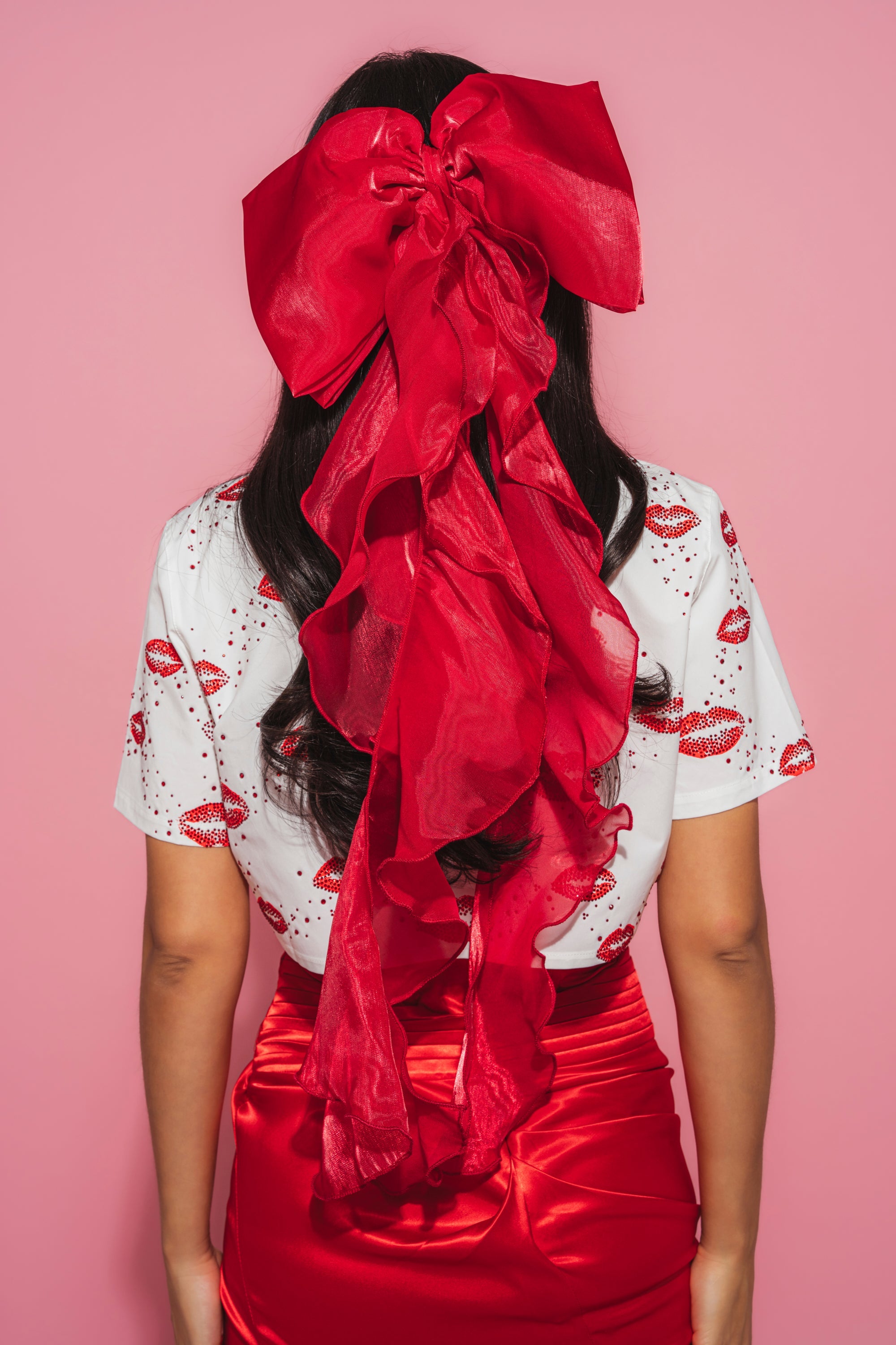 A woman with long dark hair, seen from behind, wears the HEY DOLL CHIFFON HAIR BOW in red, a white shirt with red lip prints, and a shiny red skirt, standing against a pink background. 1
