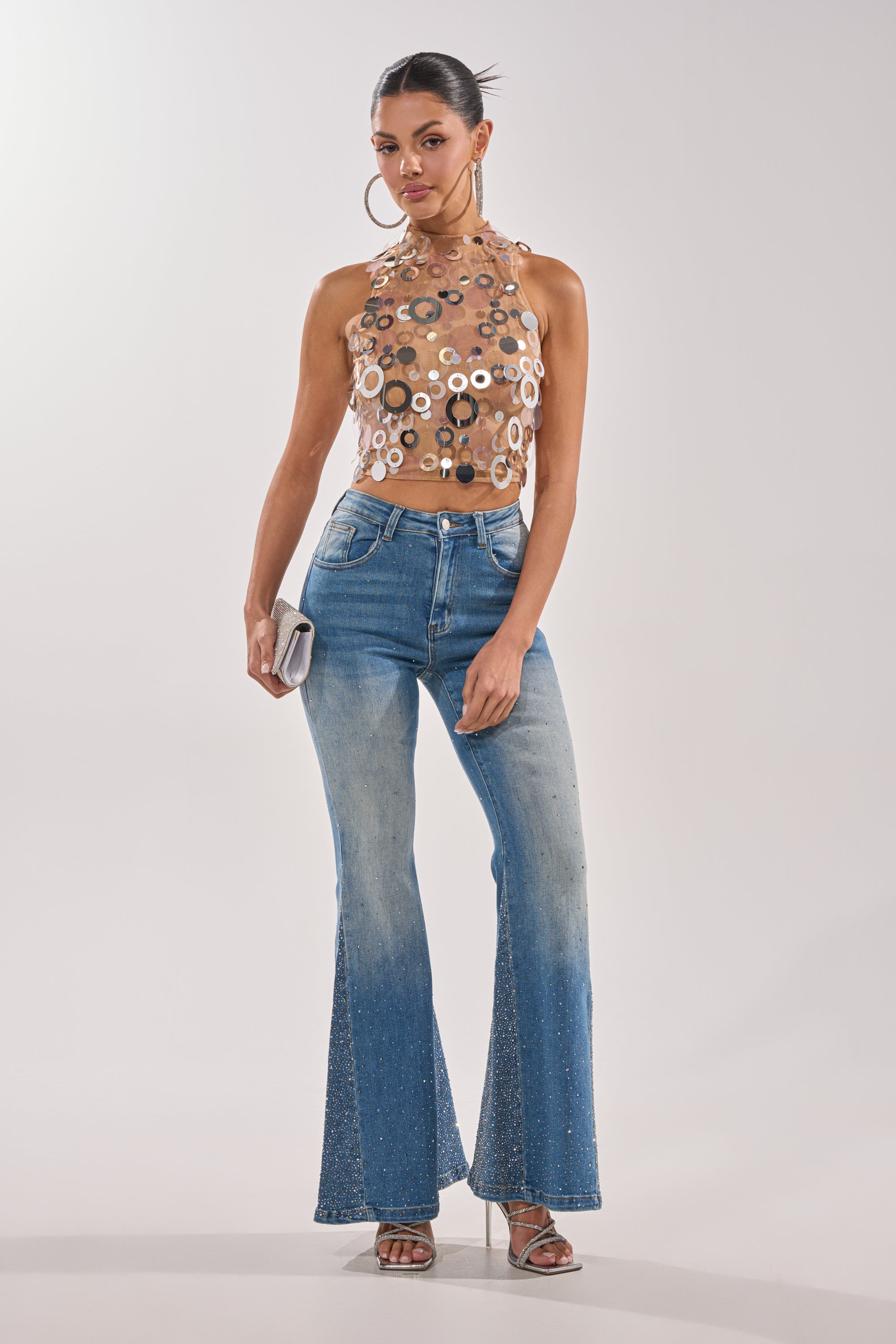 A woman stands confidently in BLING BLING FLARE DENIM, a sleeveless sheer halter top with circular accents, silver heels, large hoop earrings, and a small silver clutch. 0