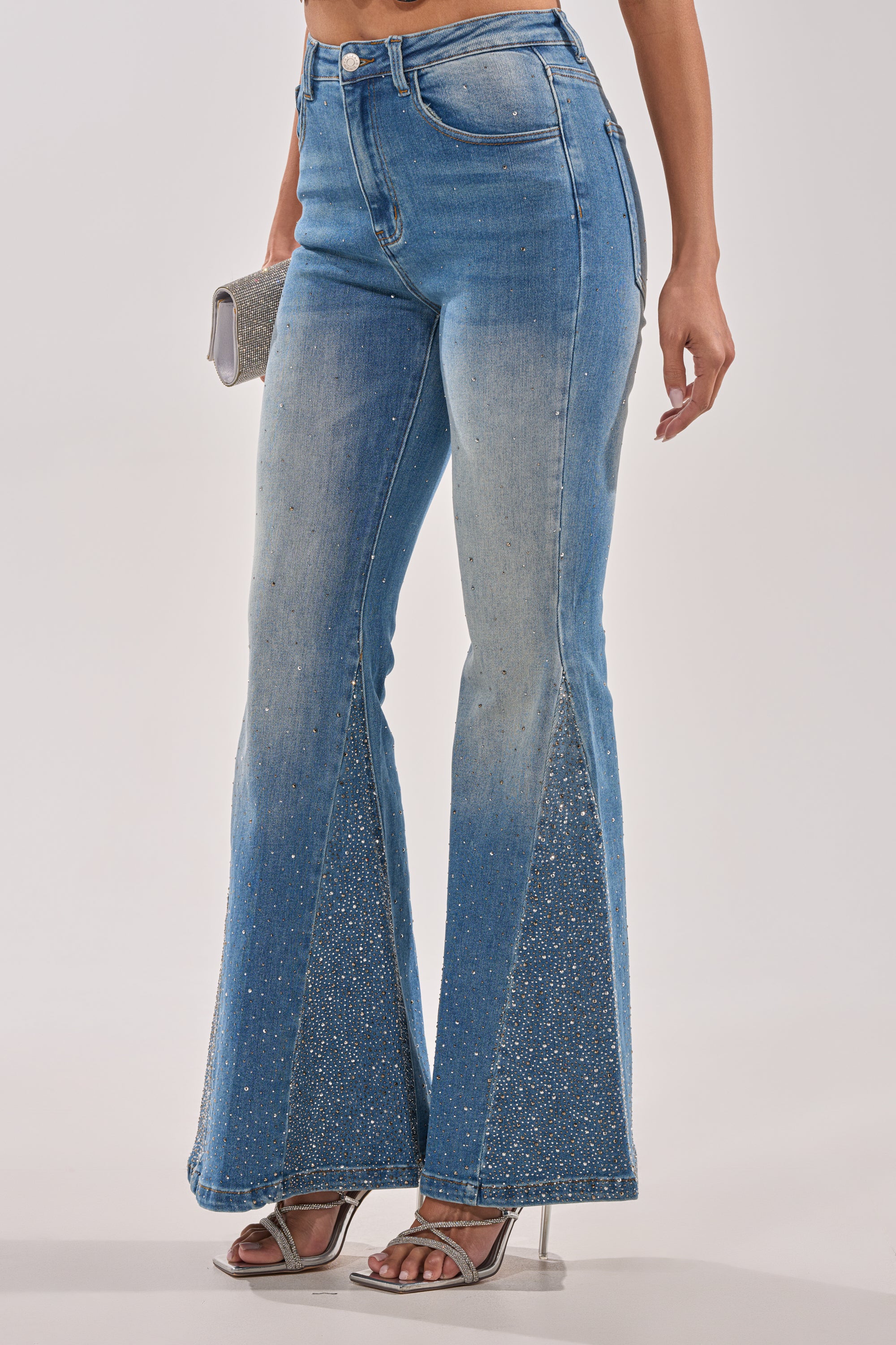 A woman models the BLING BLING FLARE DENIM with rhinestone embellishments, paired with silver high-heeled sandals and a small silver clutch; the photo is cropped from the waist down. 1