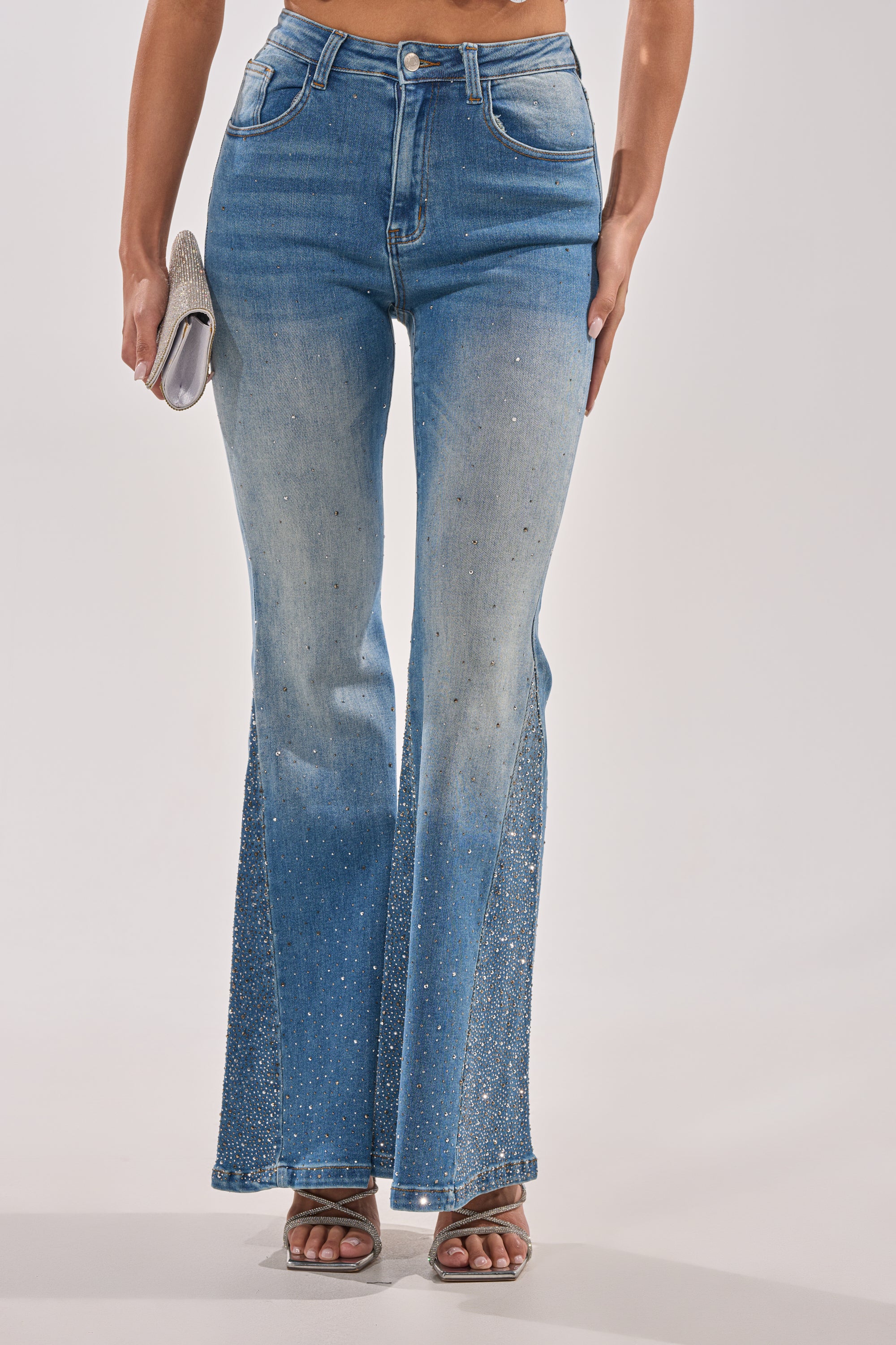 A woman wears BLING BLING FLARE DENIM with rhinestone embellishments, silver heeled sandals, and a silver clutch. The high-rise jeans add a chic twist to classic western style. The background is plain white. 2