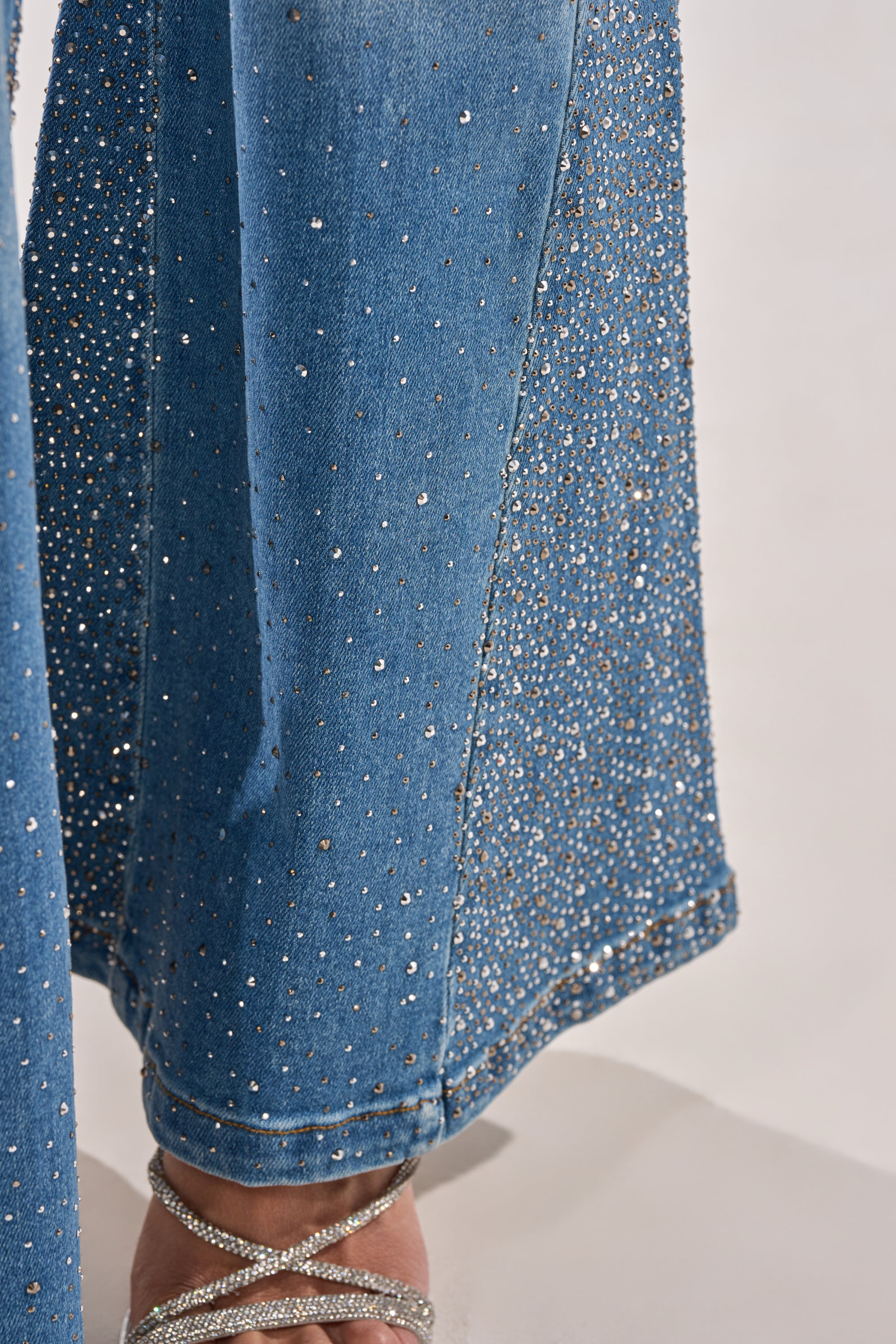 Close-up of BLING BLING FLARE DENIM, high-rise vintage-style jeans adorned with sparkling rhinestones, styled with strappy silver high-heeled sandals. 5