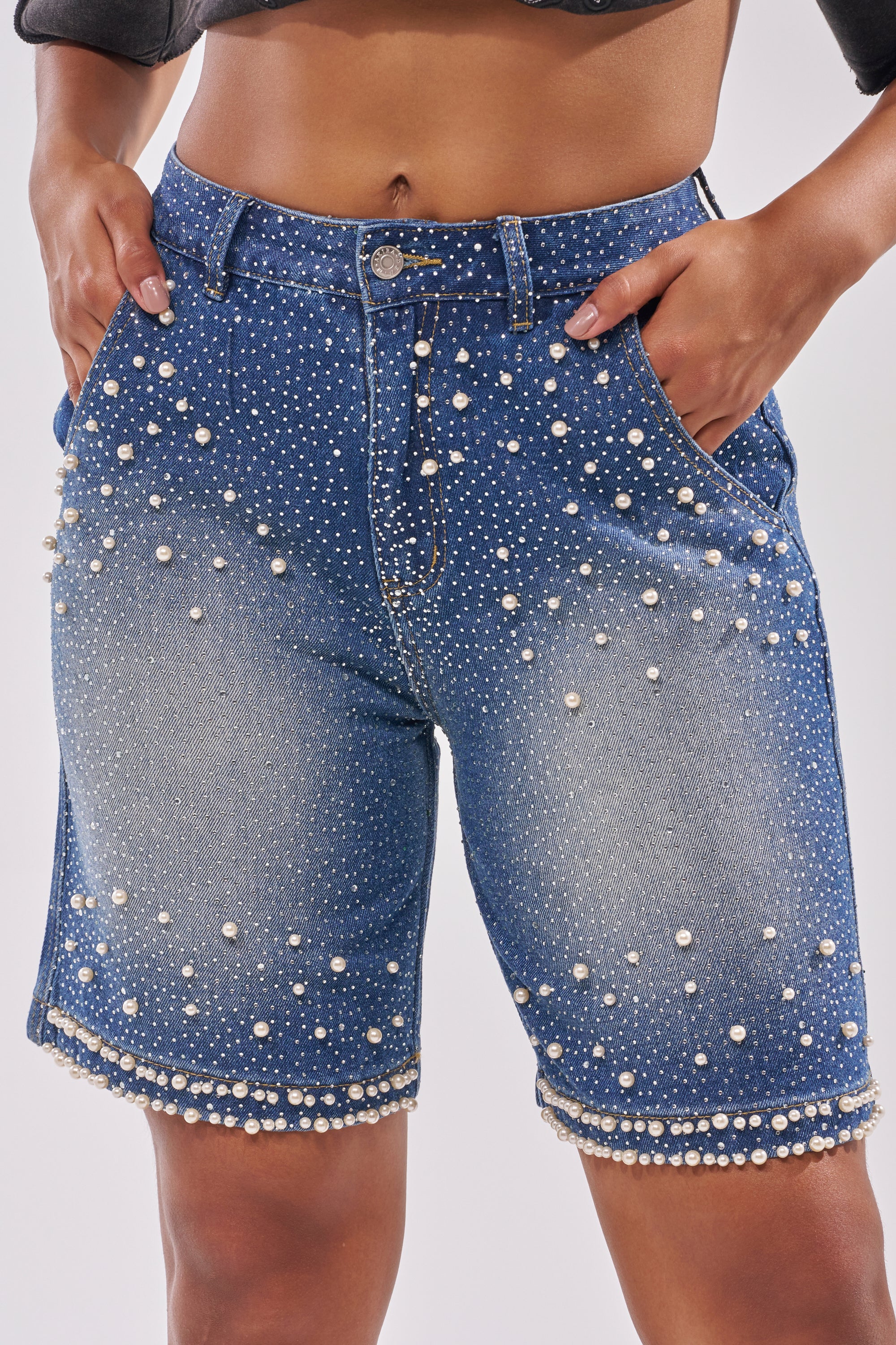 A person models the BLINGED OUT CARGO SHORTS IN MEDIUM BLUE DENIM, featuring a high waist, faded finish, and pearl and stud embellishments throughout, with hands in pockets. 1