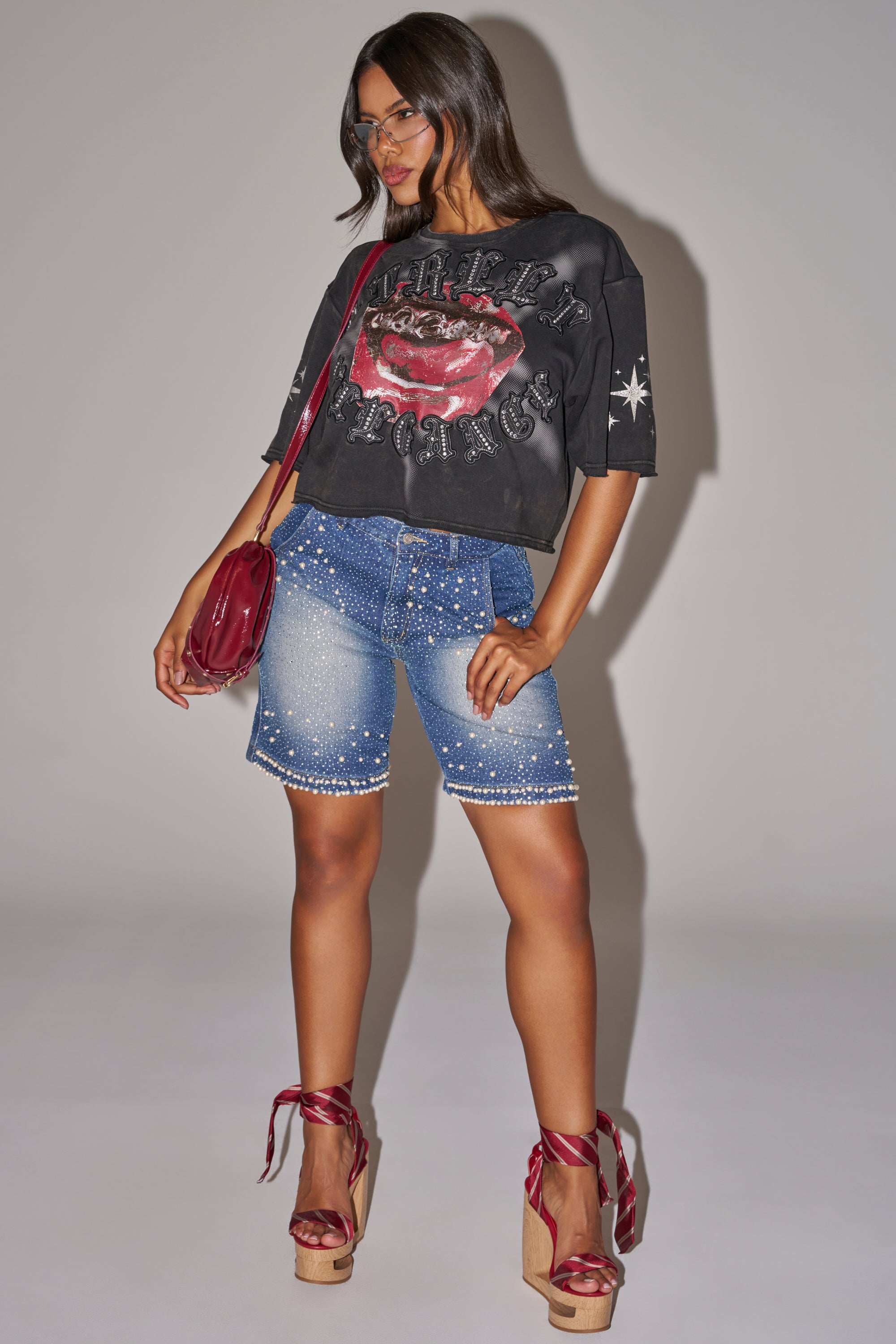 A woman poses in a loose black graphic tee, BLINGED OUT CARGO SHORTS IN MEDIUM BLUE DENIM, red satin platform heels, sunglasses, and a red shoulder bag against a plain light gray background. 6