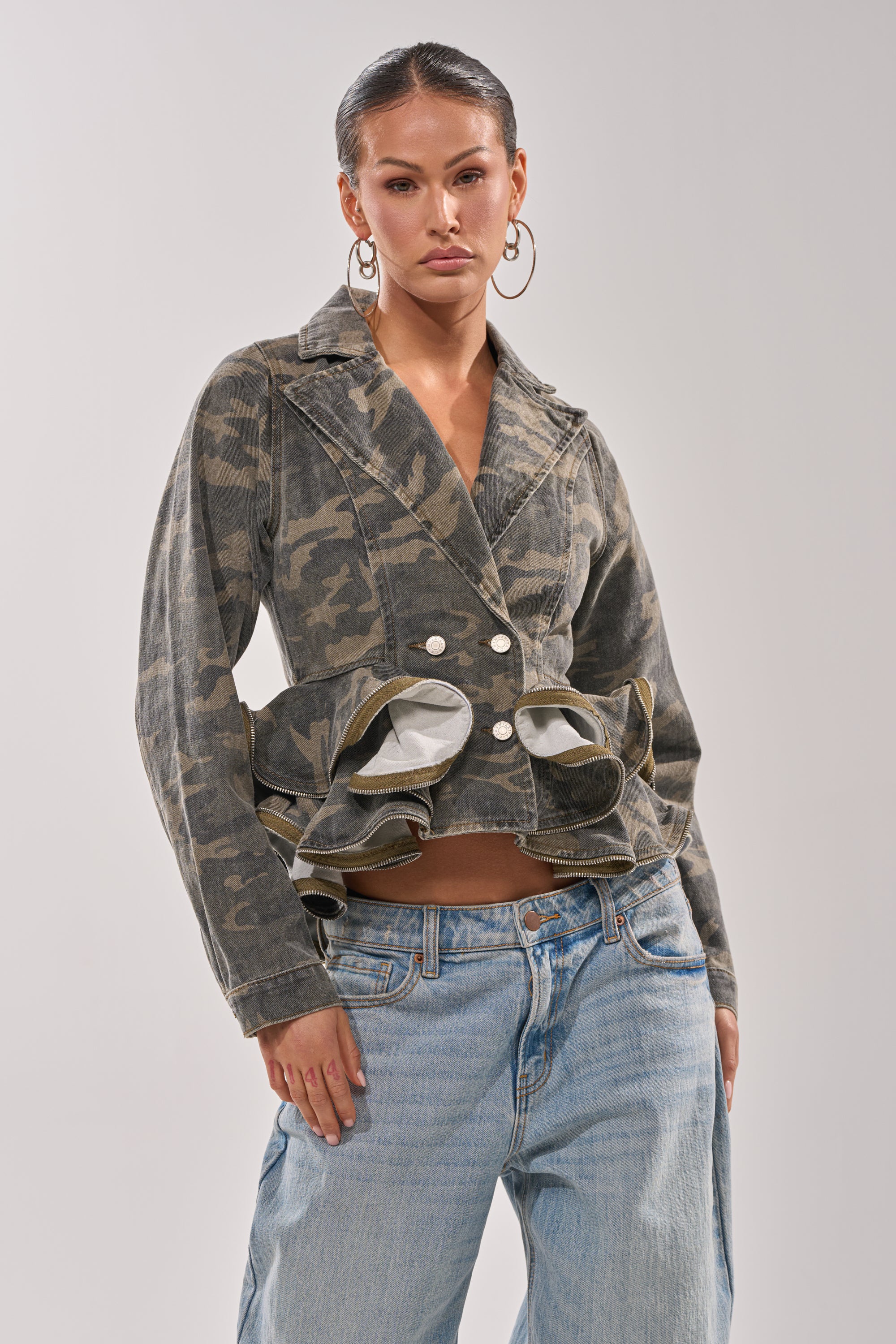 A woman in hoop earrings wears the PEPLUM CAMO JACKET with ruffled details and light blue baggy jeans, posing confidently against a plain, light background. 1
