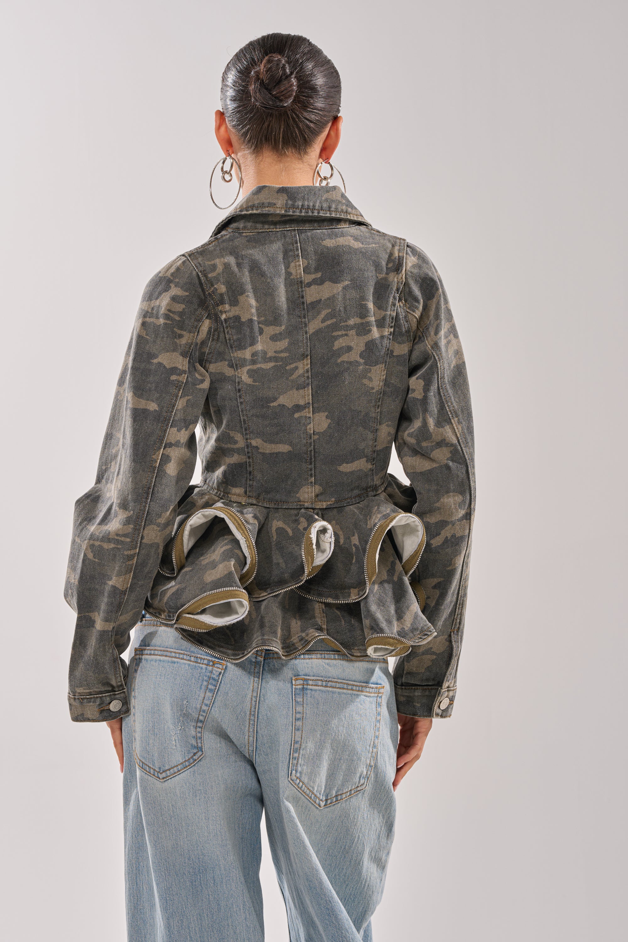 A woman models the PEPLUM CAMO JACKET with light blue jeans. Her dark hair is styled in a bun, and she wears large hoop earrings, creating an effortless street style look against a plain light background. 4