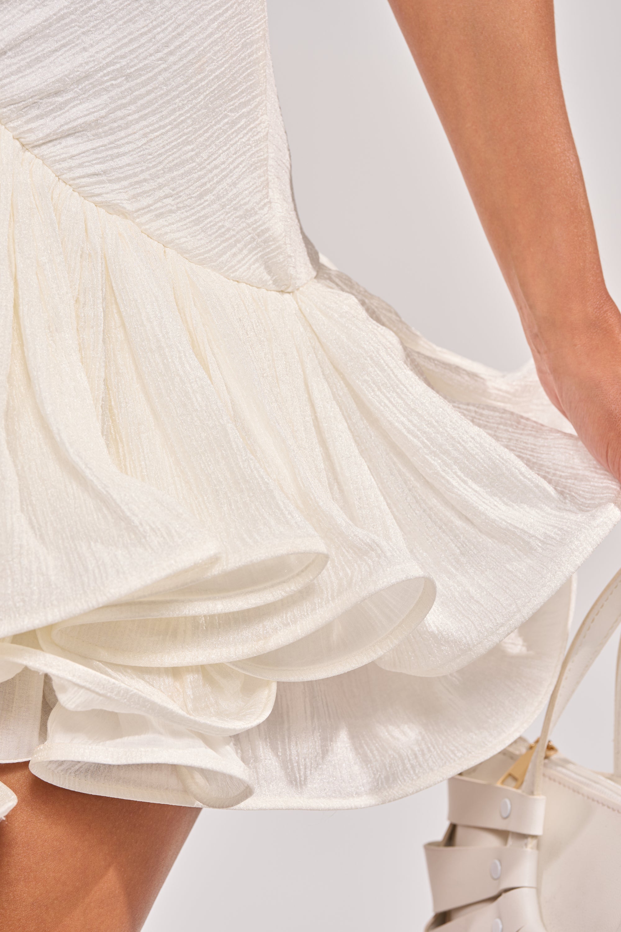 Close-up of a person wearing the WILD HEART HALTER MINI DRESS in off-white with a tiered ruffle hem, holding a white handbag. The focus is on the flowing fabric and their arm against a soft, minimal background. 9