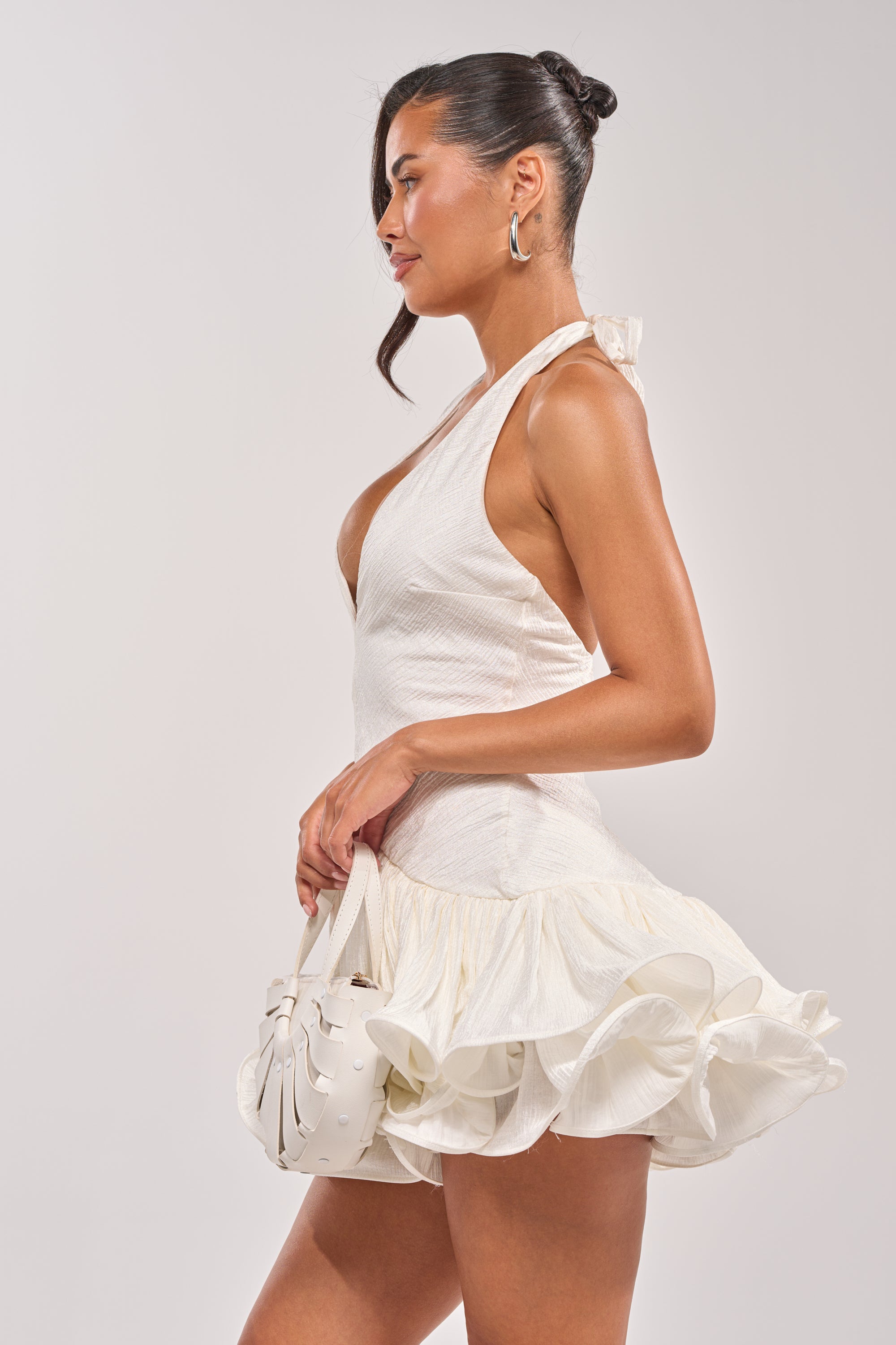 A woman stands in profile wearing the WILD HEART HALTER MINI DRESS and holding a small white handbag. Her hair is in a sleek bun, hoop earrings shine, and she poses elegantly against a plain light background. 4
