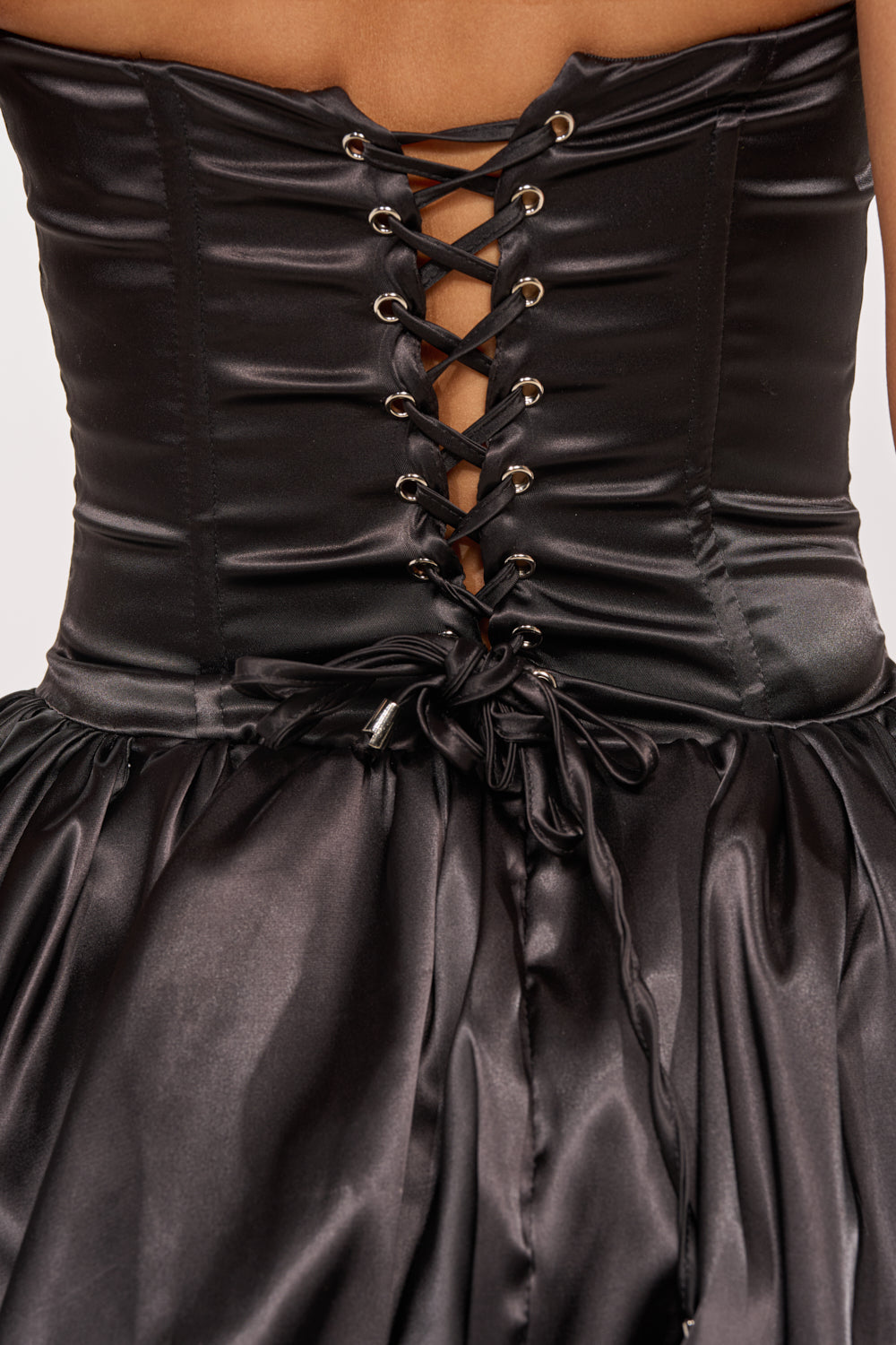Close-up of the back of the CRUSH ON YOU STRAPLESS CUT OUT MINI DRESS in black satin, showcasing a lace-up design with silver eyelets, a tied waist bow, fitted bodice, and voluminous bubble hem skirt. 10