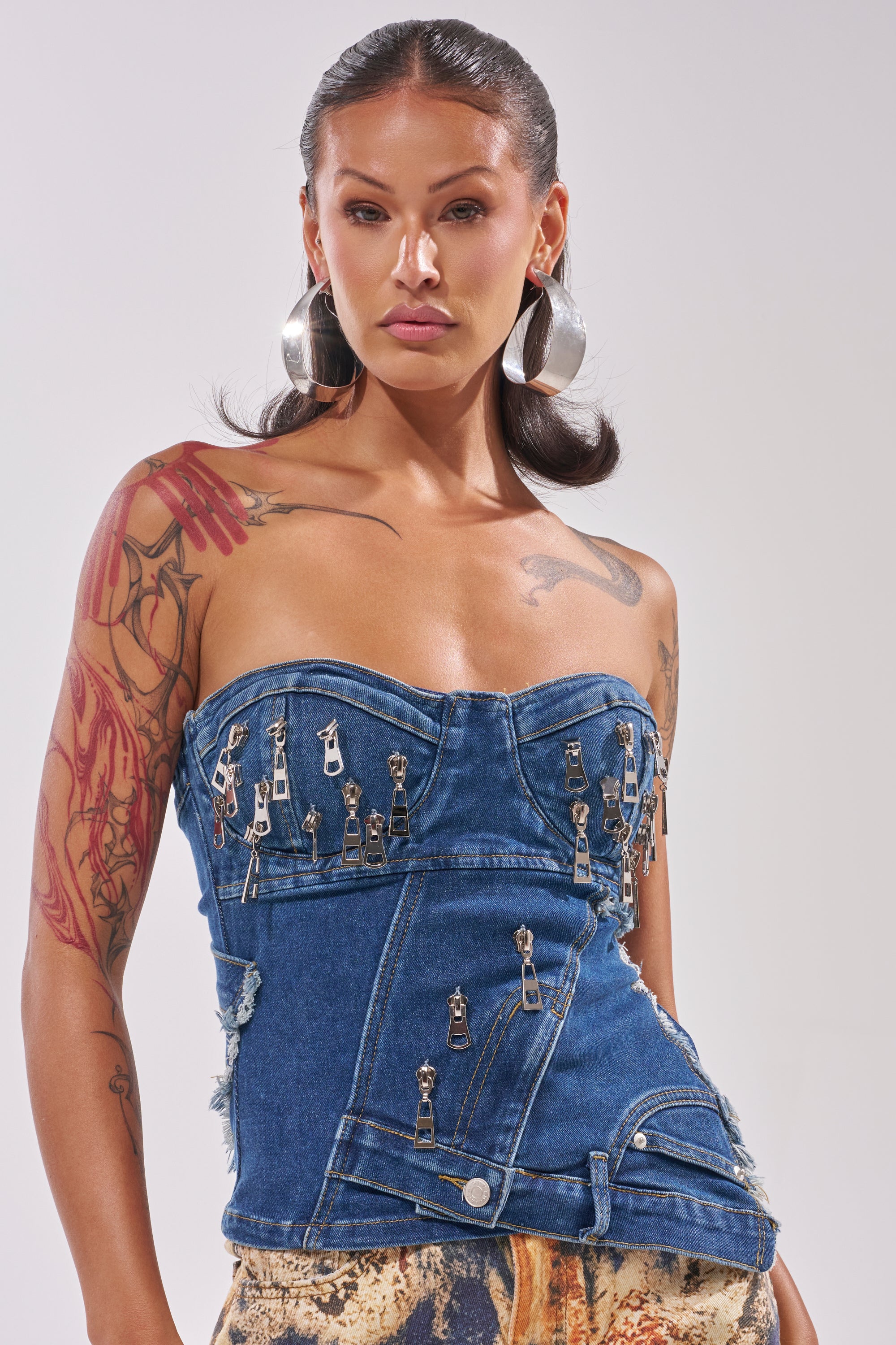 A person with long dark hair, hoop earrings, and tattoos wears the CHAINED OUT CORSET—a strapless indigo denim top with a sweetheart neckline and multiple zippers—paired with a printed skirt, against a plain light background. 0