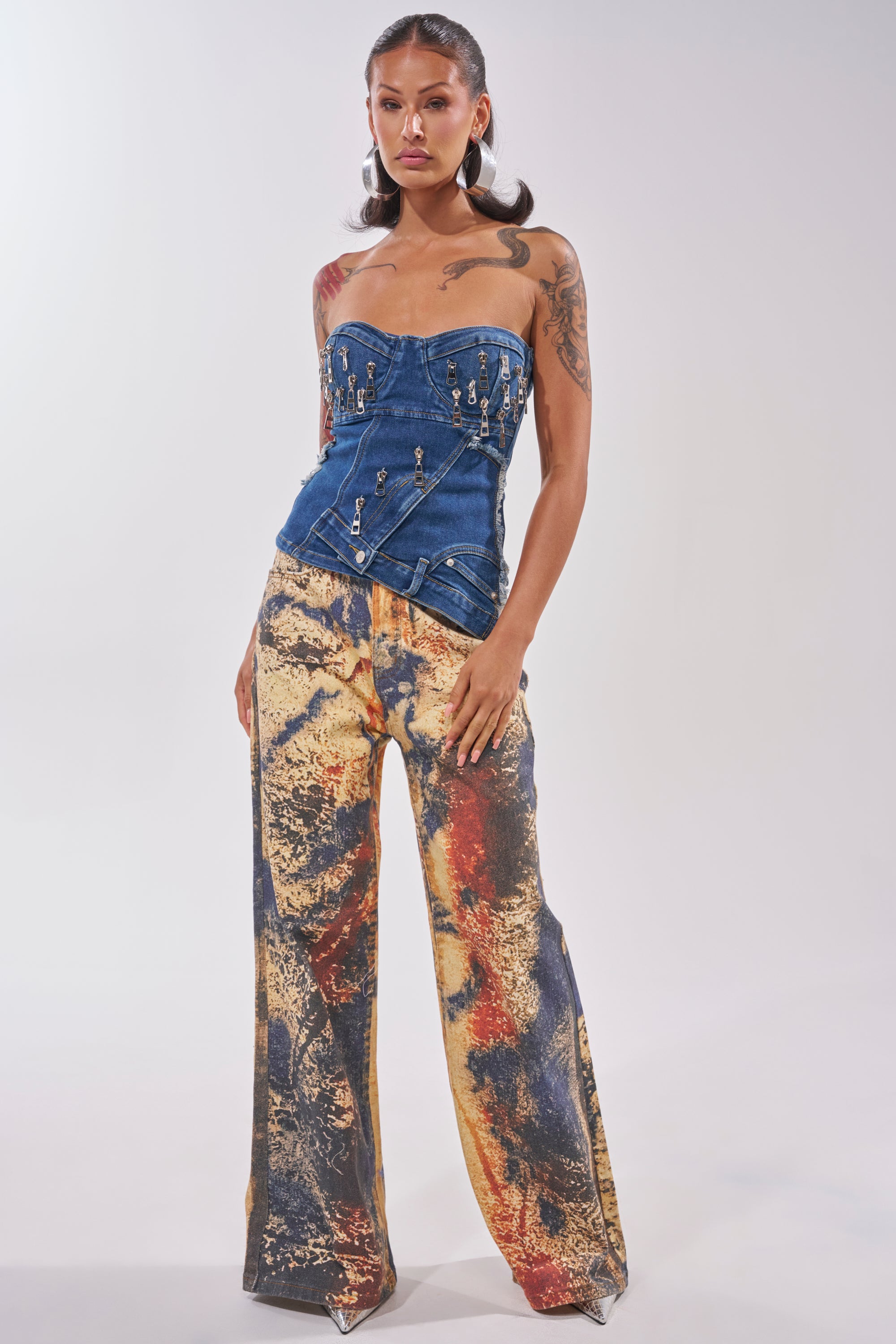 A woman models the CHAINED OUT CORSET, a strapless indigo denim top with metallic details, paired with wide-leg pants showcasing a bold abstract yellow, red, and black pattern against a plain background. 4