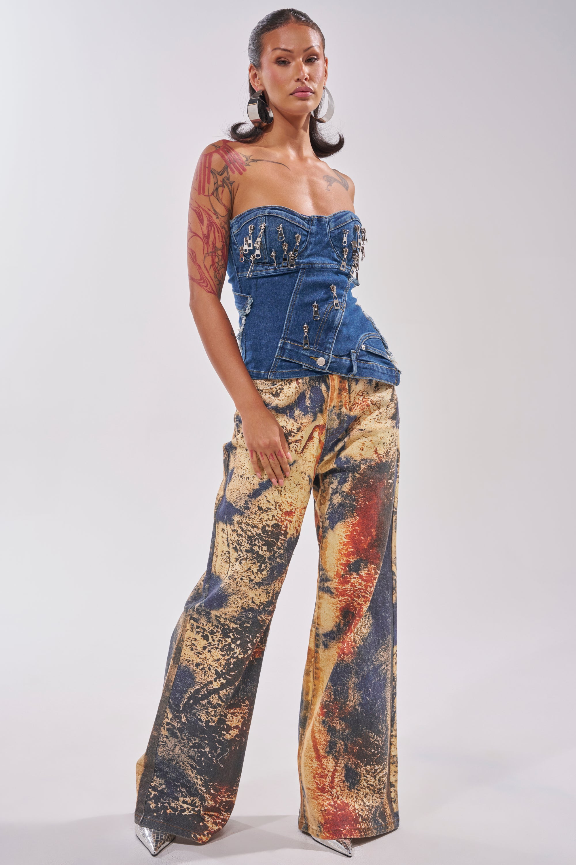 Wearing the CHAINED OUT CORSET, a structured denim piece with metal studs and zippers, they pair it with wide-leg trousers in yellow, orange, and black abstract patterns. Tattoos are visible on one arm. 5