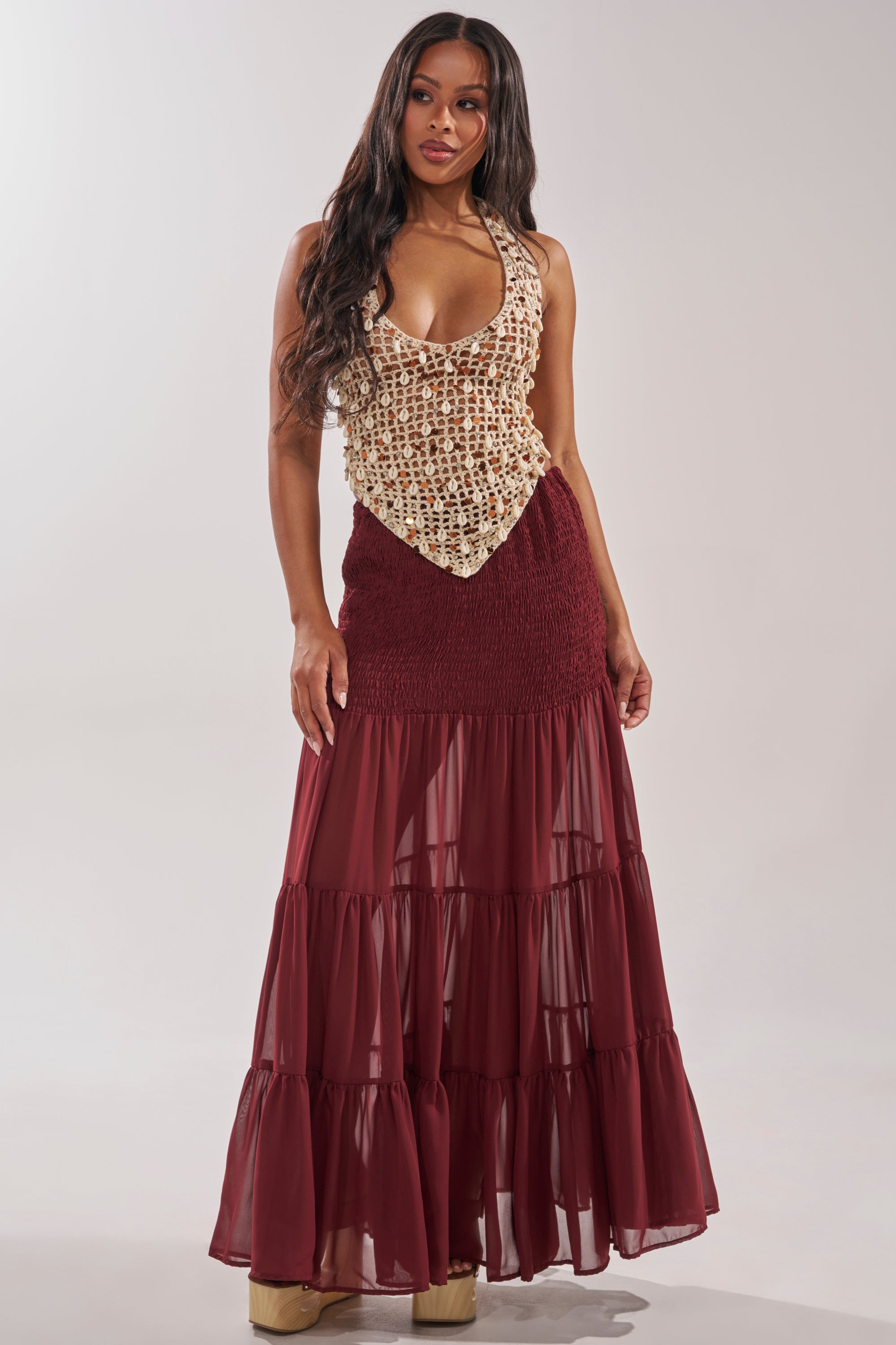 A woman with long, wavy hair poses against a plain background in a beige crochet top and the DRIFTING AWAY MAXI SKIRT—a maroon, tiered maxi skirt with sheer layers—paired with platform shoes. 0