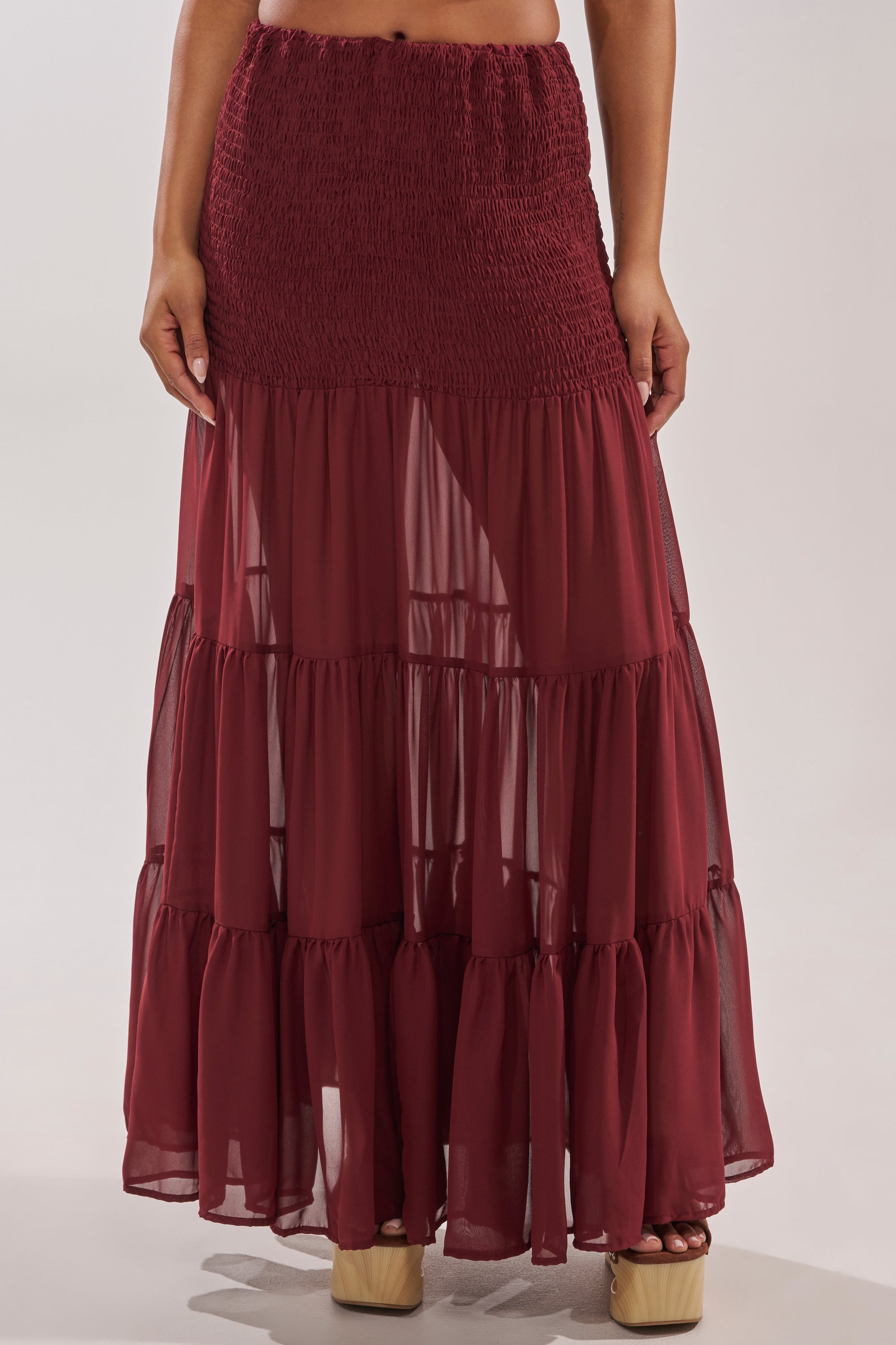 A person wearing the DRIFTING AWAY MAXI SKIRT, a smocked high-waist, tiered maxi skirt in sheer burgundy, paired with beige platform sandals, shown from the waist down against a plain background. 1