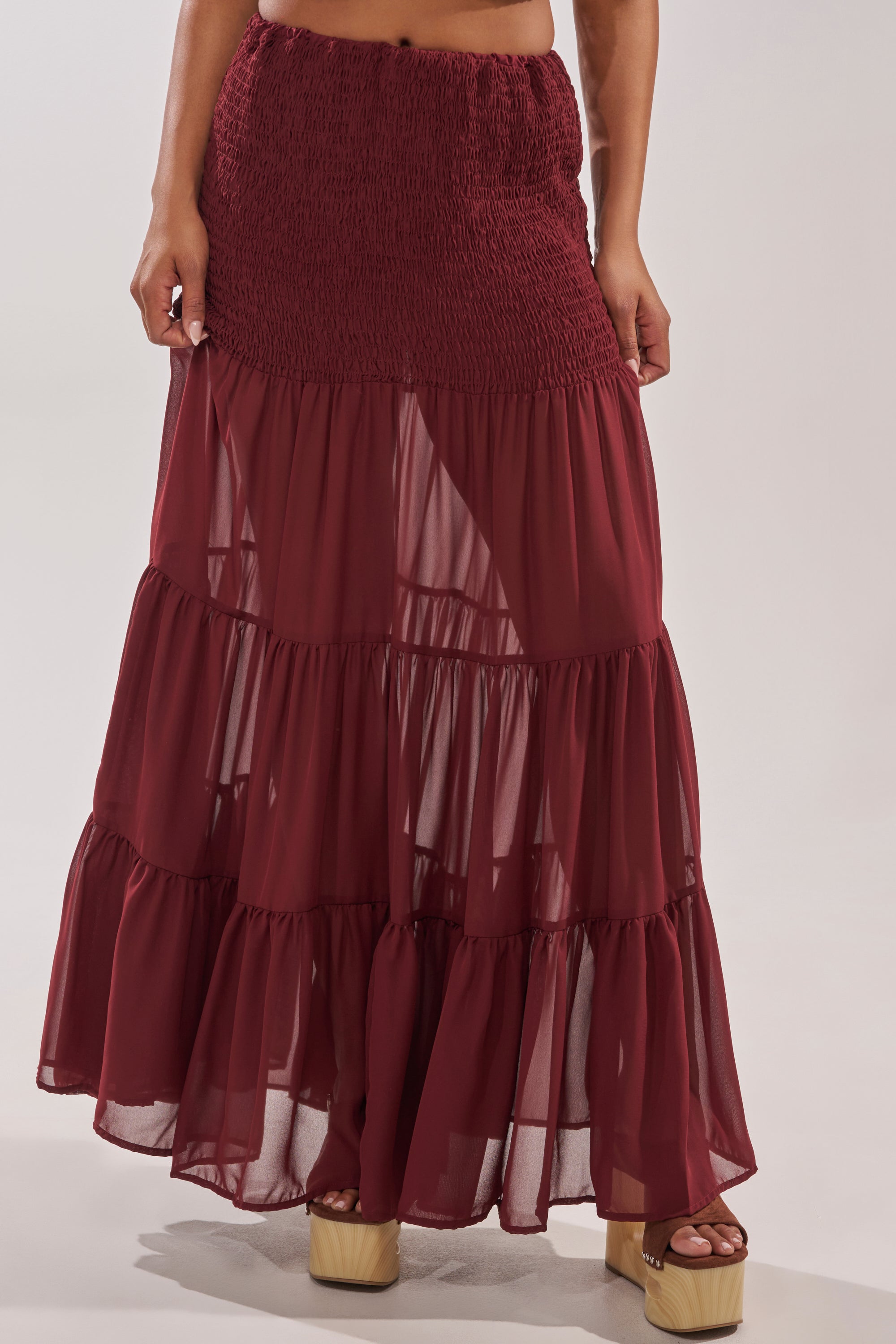 Someone wearing the DRIFTING AWAY MAXI SKIRT, a maroon maxi with a smocked high-waist and sheer, flowy tiers, styled with beige platform sandals. 2