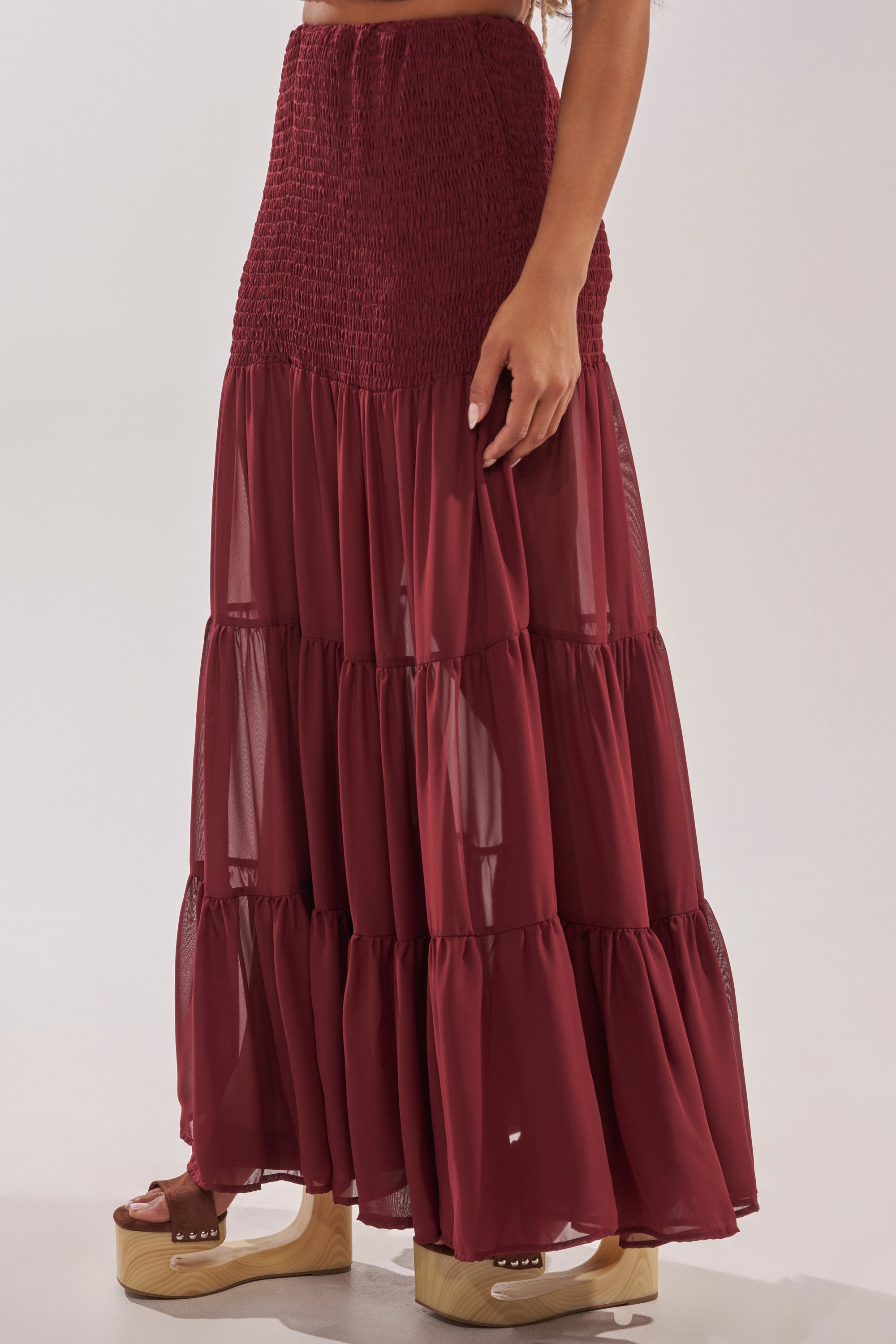 Wearing the DRIFTING AWAY MAXI SKIRT, a person showcases its burgundy sheer tiers and smocked high-waist with tan platform sandals, standing against a plain light background. 3