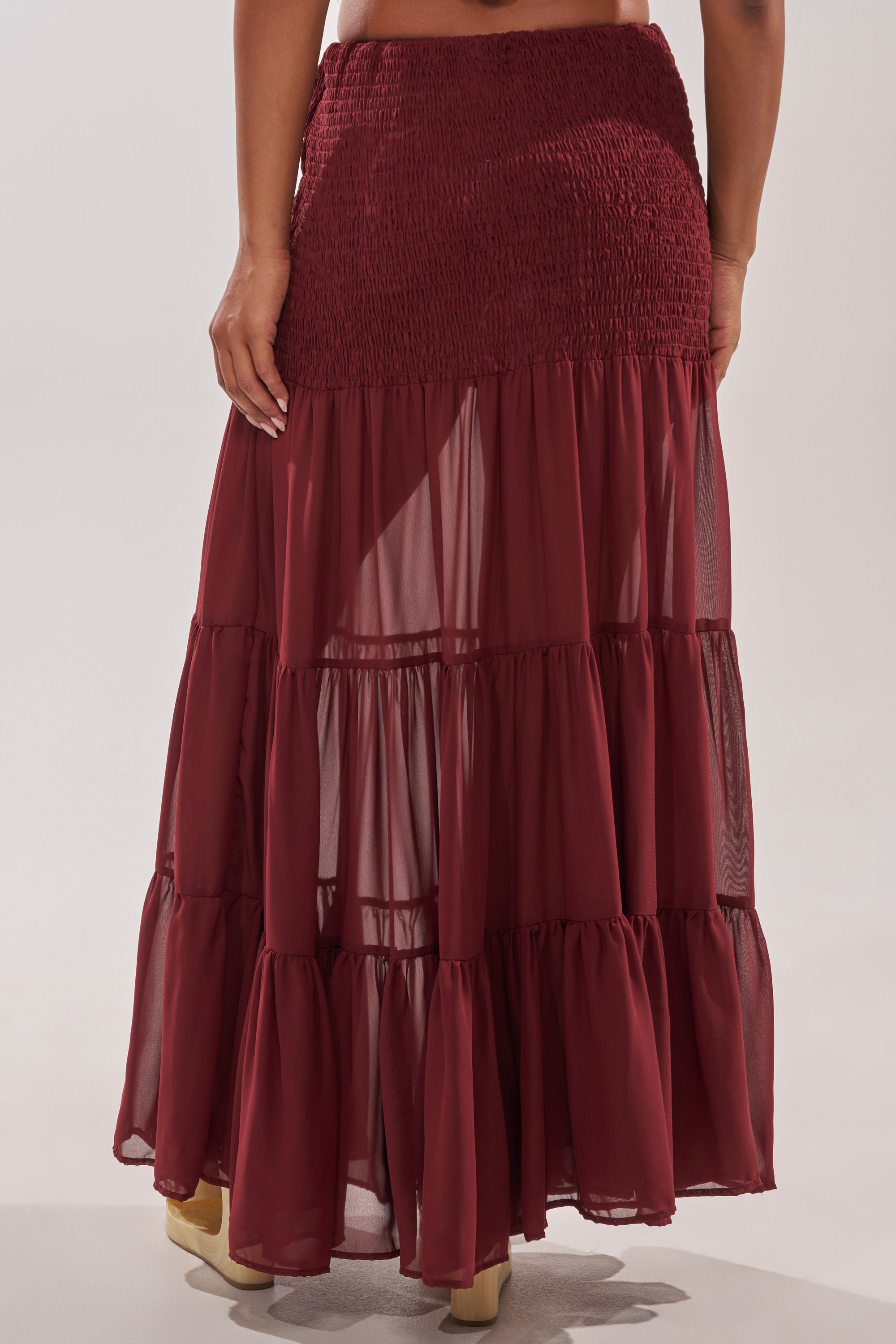 Wearing the DRIFTING AWAY MAXI SKIRT, a tiered burgundy maxi with a smocked high-waist and sheer fabric, this person stands with their back to the camera. 4